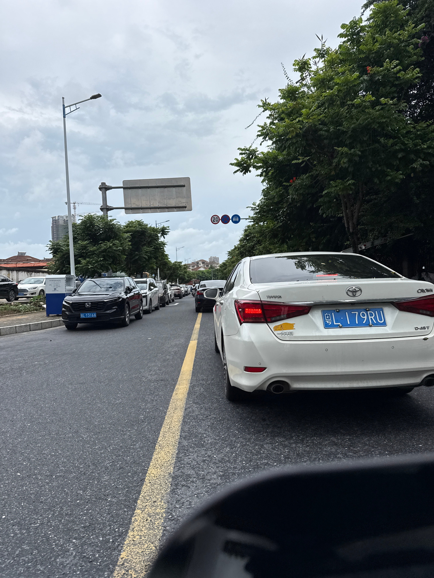 The image captures a street scene in Huizhou, China, characterized by a traffic jam. The foreground is dominated by the rear of a white Toyota Crown sedan, displaying a blue license plate that reads "L·179RU". A yellow dividing line on the asphalt road runs from the bottom right to the middle of the frame. To the left of the white sedan, a black Honda SUV is visible, its license plate partially obscured but showing "BL·Y316A". Multiple other vehicles are lined up behind these two, indicating a significant traffic backup. Along the left side of the road, a row of trees forms a green barrier, behind which are buildings and construction cranes, suggesting an urban environment. Overhead, a cloudy sky looms. Traffic signs, including a speed limit sign with "20" and a blue directional sign, are present on the right side of the road, above the trees.