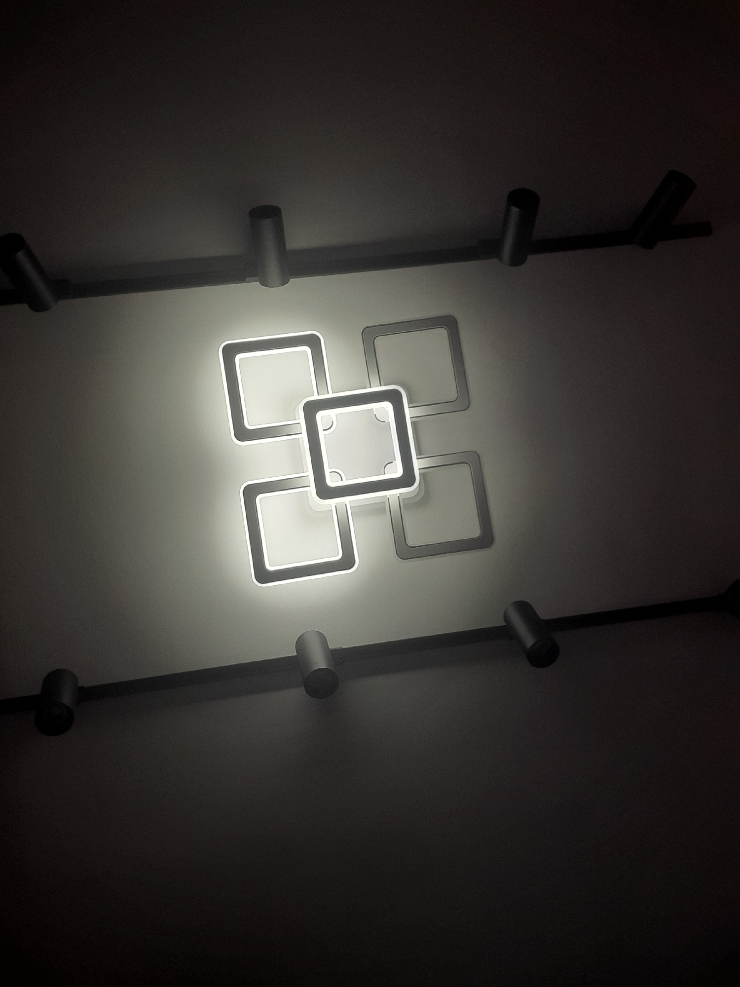 A modern ceiling light fixture composed of interlocking square frames is illuminated against a dark ceiling. The fixture is mounted on a black linear track that extends across the ceiling, with several cylindrical spotlights attached to it. The square frames of the light fixture are outlined with bright LED lighting, creating a central, brighter square in the middle of the arrangement. The background is a smooth, dimly lit ceiling, with shadows cast by the light fixture and the spotlights. There are no people or any other identifiable objects in the scene.