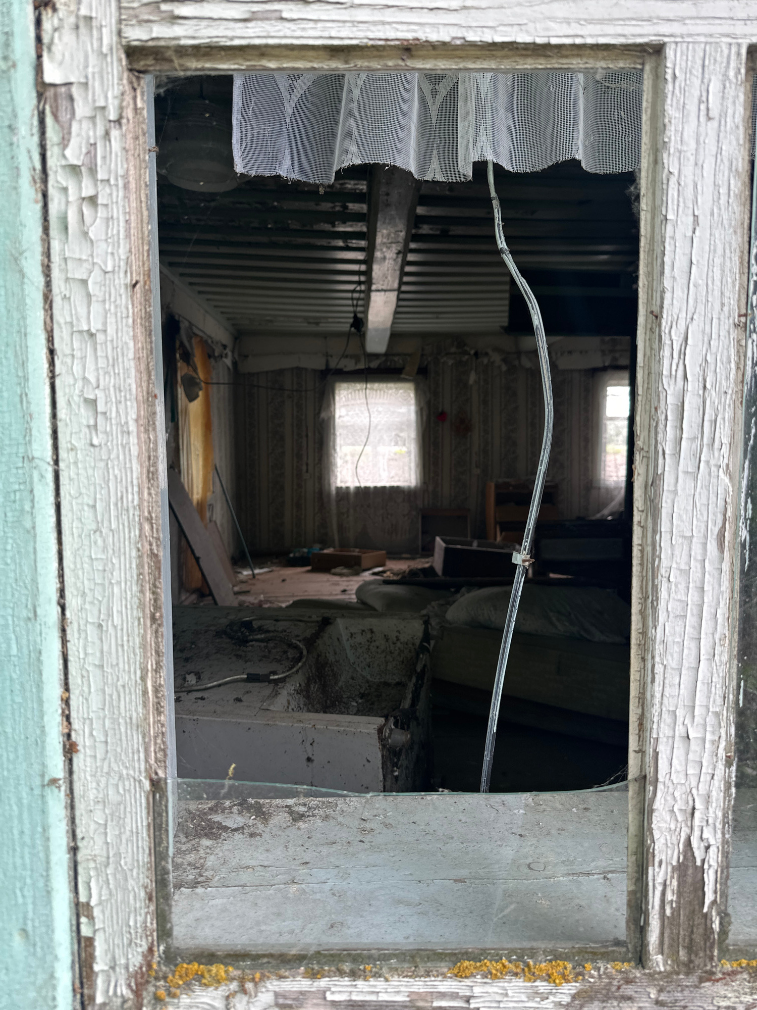 The image shows a view through a broken window into a dilapidated room. The window frame is old and weathered, with peeling paint. The room inside is sparsely furnished and appears abandoned. There is a mattress visible on the floor, along with other debris and household items. The walls are covered in patterned wallpaper, and the ceiling is exposed with visible beams and wiring. A sheer curtain hangs across the top of the window. The overall impression is one of decay and neglect. The image location is not specified beyond being in the Відзаўскі сельскі Савет, Belarus.