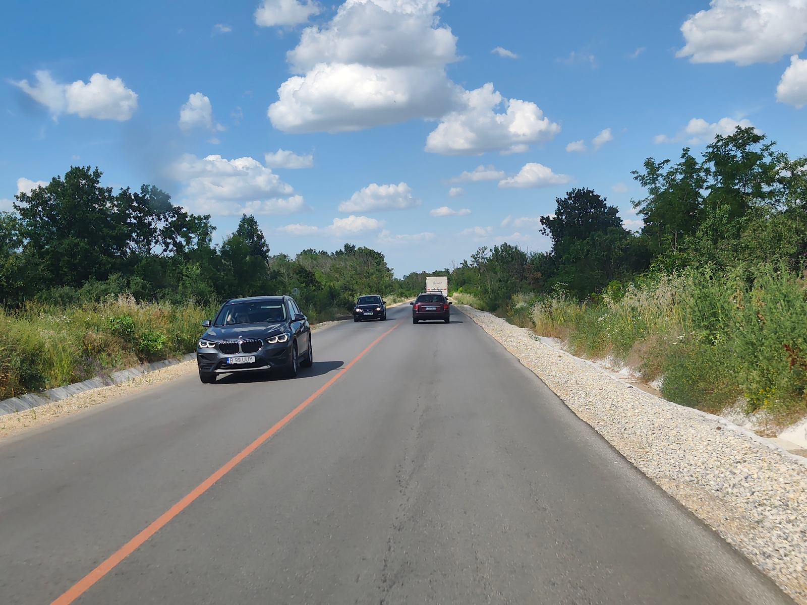 A forward-facing, eye-level perspective shot captures a two-lane asphalt road with a bright orange lane divider. On the left side of the road, a dark-colored BMW SUV, license plate B 99 UUY, is in the foreground. Further down the road, a dark car and a white truck are visible. The right side of the road features a gravel shoulder bordered by lush green vegetation and trees. The sky is a vibrant blue, dotted with fluffy white clouds. The scene appears to be a rural setting, possibly on the outskirts of Aglen, Bulgaria.
