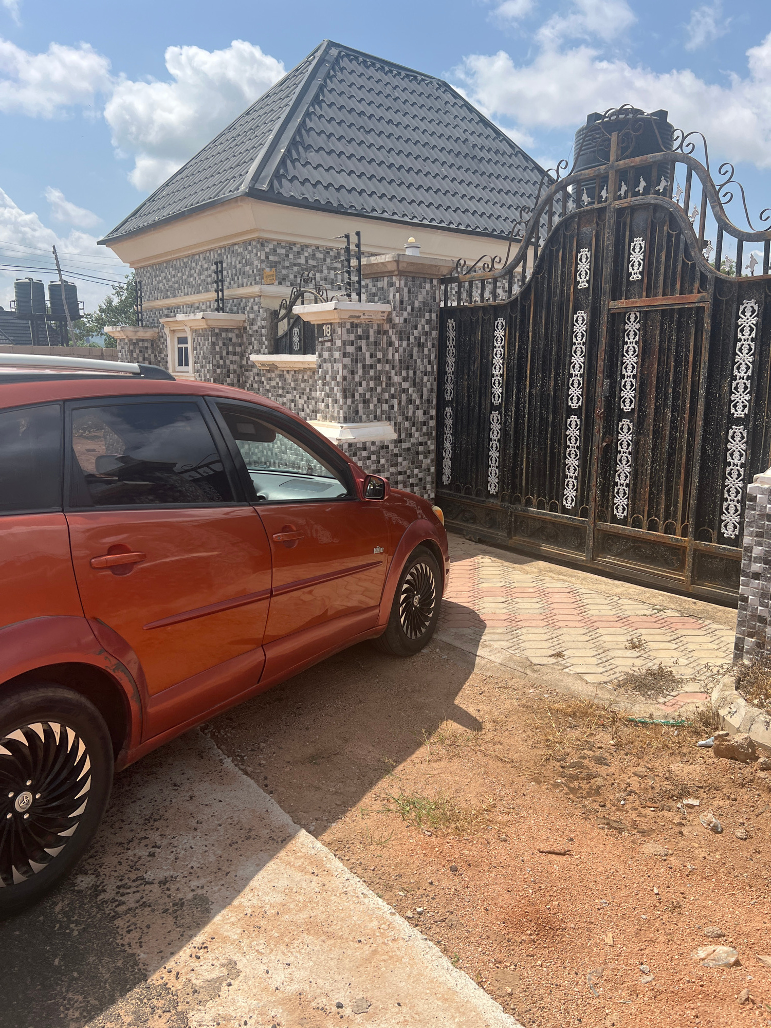 An orange car is parked on a dirt and paved driveway in front of a large, ornate black metal gate. Behind the gate is a building with a grey, tiled roof and a facade of grey and white patterned tiles. The gate is decorated with white scrollwork. In the background, two large water tanks are visible, along with a utility pole. The sky is blue with scattered white clouds. The scene is lit by bright sunlight, casting shadows.