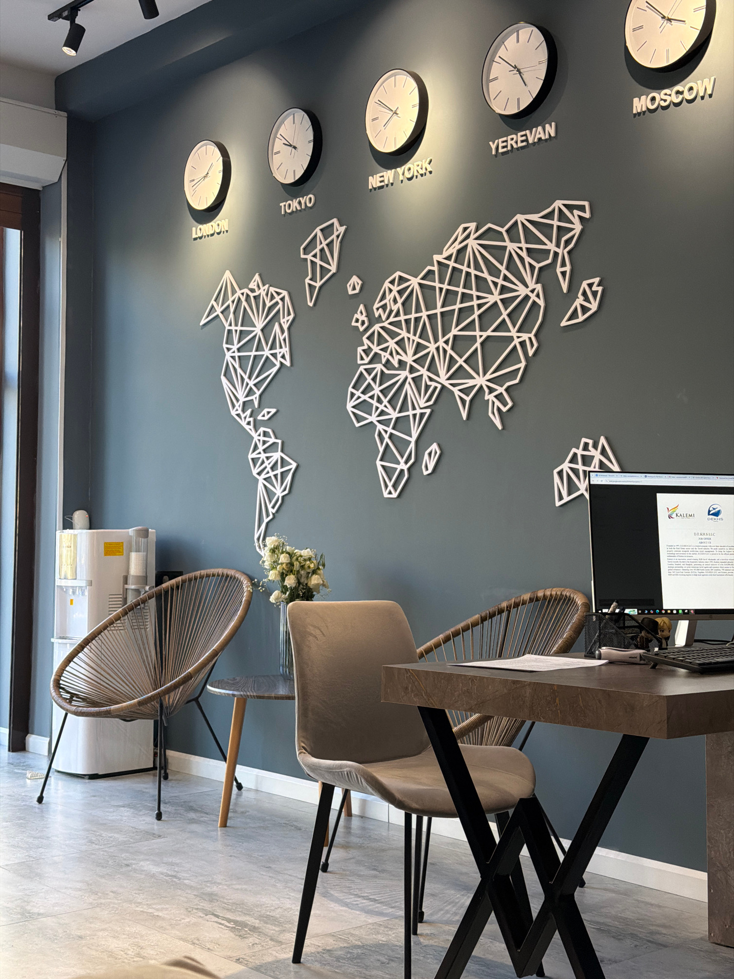 The image shows a modern office interior with a dark blue wall adorned with a white geometric world map and five clocks, each displaying a different city's time: London, Tokyo, New York, Yerevan, and Moscow. A water cooler stands to the left, next to a window. Two chairs are visible: one with a woven design and the other a plush grey armchair. A dark wood desk with a computer and office supplies sits in the foreground. The overall impression is one of a stylish and organized workspace, likely in a location that deals with international business or travel, such as an office in Yerevan, Armenia.