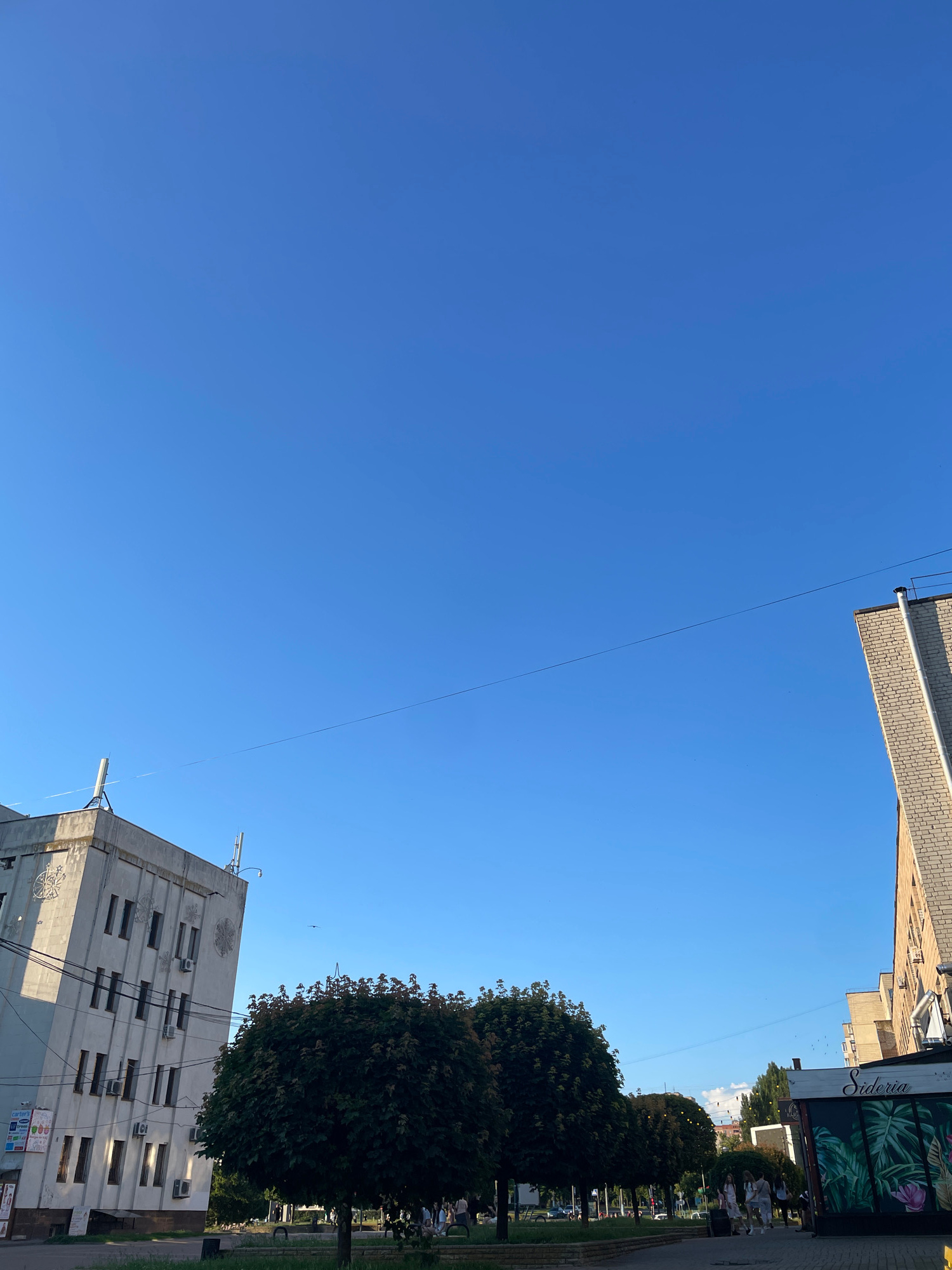 This is a ground-level, full-frame, eye-level, outdoor, daylight, slightly low-angle shot looking up at the sky and buildings in Brovary, Ukraine. The sky is clear and blue. On the left, a light-colored, multi-story building with many windows and architectural embellishments is visible. In the center, two large, dark green, rounded trees are prominent. To the right, another building constructed of light-colored bricks is seen. In the foreground, a paved walkway is present, with several people walking. A sign for "Sideria" with a tropical leaf motif is visible on a storefront to the right. The image captures a section of a city street with buildings, trees, and pedestrians under a bright blue sky.