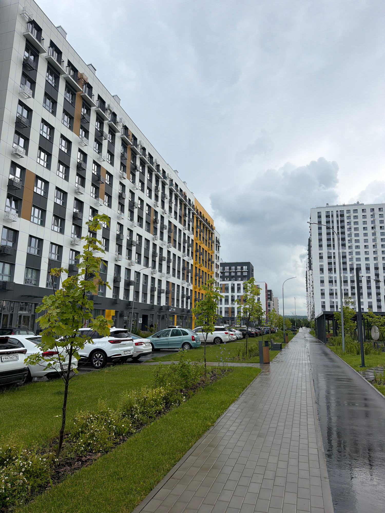 The image depicts a modern residential complex under a cloudy sky. On the left, a large, multi-story building with a white, gray, and yellow facade dominates the frame. It features numerous windows and balconies with dark railings. To the right of this building, a row of parked cars is visible, lined up along a green lawn that is bordered by a paved walkway. Several young trees with green foliage are interspersed along the walkway.
Further to the right, another tall, white building with a grid-like pattern of windows stands prominently. This building also has a paved walkway running alongside it, which is wet and reflects the overcast sky, suggesting recent rain. The walkway extends into the distance, flanked by more greenery and trees. In the background, other buildings of similar modern architecture can be seen, creating a sense of a planned urban development. The overall impression is of a clean and orderly, albeit somewhat muted, urban environment.