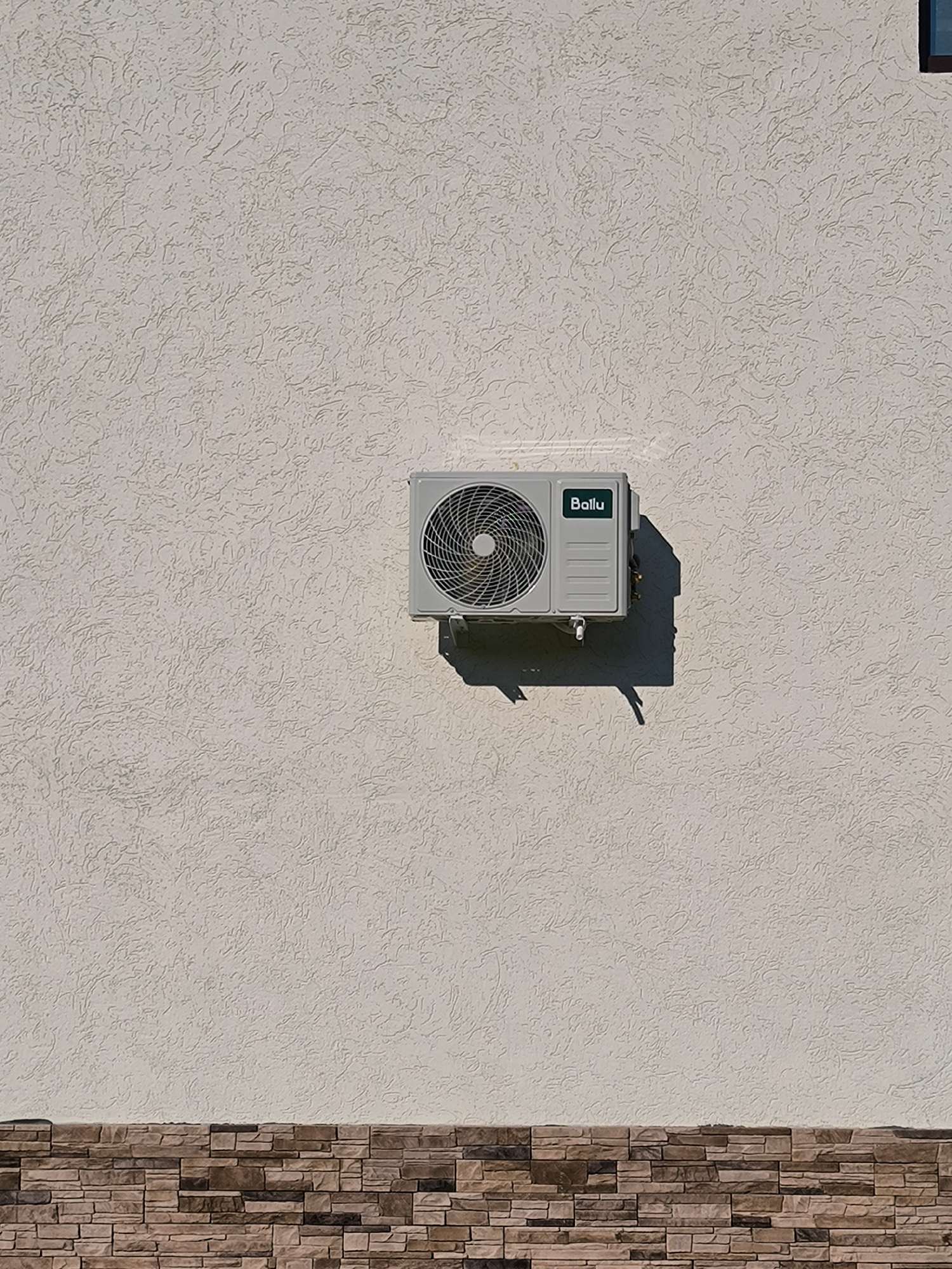 A light-colored, textured wall is the backdrop for a mounted air conditioner unit. The wall has an irregular, stucco-like finish with subtle shadows cast across its surface, indicating it is illuminated by sunlight. The air conditioner is a rectangular, light gray unit with a circular fan grill on the left side and a green rectangular panel on the right. Visible text on the green panel reads "Ballu." The unit is mounted to the wall with a dark bracket that casts a sharp shadow. Below the wall, a section of stone veneer is visible, with rectangular stones in shades of brown and gray arranged in a horizontal pattern. The lighting suggests it is daytime, likely sunny, and the scene is outdoors. There are no people visible, and the overall impression is one of a functional building exterior.