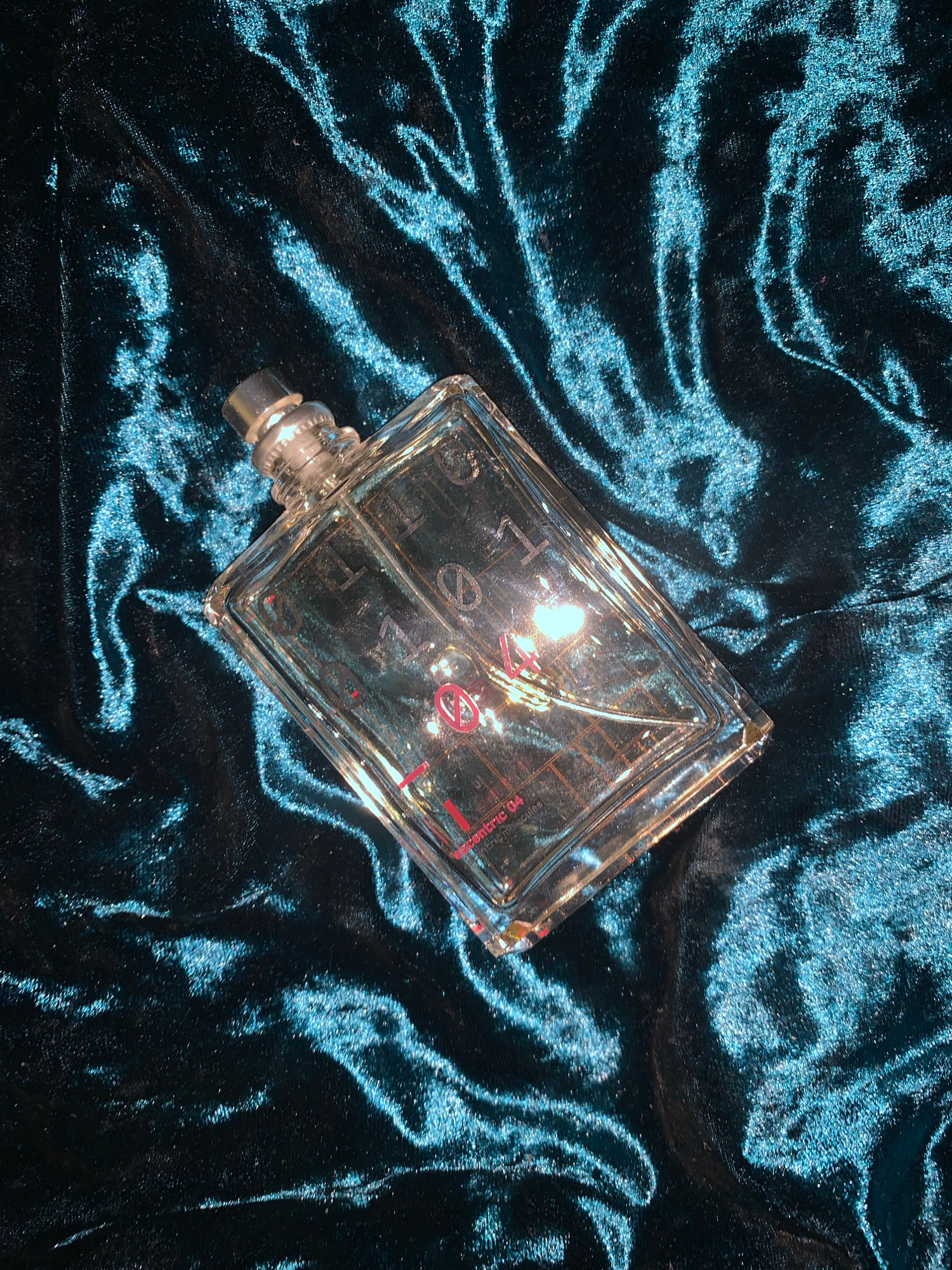 A clear glass rectangular perfume bottle with a silver cap rests on a dark teal velvet fabric. The fabric has a rippled texture that catches the light, creating a luminous effect. The perfume bottle is positioned diagonally across the frame, with the cap at the top left and the bottom right corner pointing towards the lower right. The bottle appears to be partially filled with a clear liquid, and red text is visible through the glass, reading "ESCENTRIC 04" and "MOLECULES". The lighting suggests a close-up shot, emphasizing the texture of the fabric and the details of the perfume bottle. There are no people or other objects present in the image, and the location is not specified.