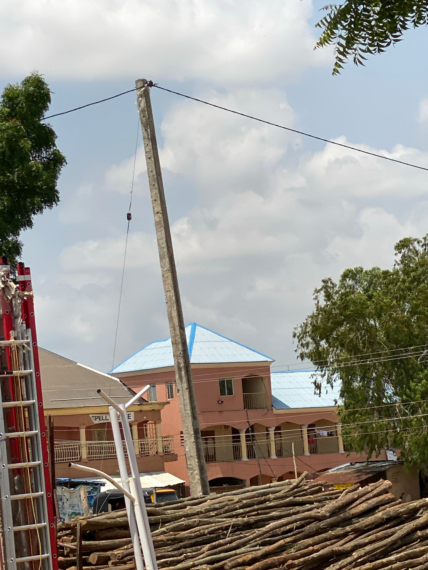 This image captures an outdoor scene with construction or utility work in progress. The central focus is a long, concrete pole, likely a utility pole, suspended in the air by wires. It appears to be in the process of being erected or lowered.
In the background, a multi-story building with a pinkish-brown facade and balconies is visible. Parts of the building are covered with a light blue tarp, suggesting ongoing construction or repair. To the right of the building, a large stack of rough-hewn wooden poles or logs stands vertically.
In the foreground, a red ladder, characteristic of emergency services or construction equipment, is positioned horizontally. A portion of a tree with green foliage is visible in the lower left corner, and another tree canopy is seen in the upper part of the image.
The sky is partly cloudy with a mix of white clouds and patches of blue sky, indicating daylight, likely mid-morning or afternoon. The overall atmosphere suggests an active work site. The location is identified as Jalingo, Nigeria.
There is some visible text on the building in the background, appearing as "SPELL" in white letters on a dark background. The scene conveys a sense of work being done, possibly involving the installation or maintenance of electrical or telecommunication lines.