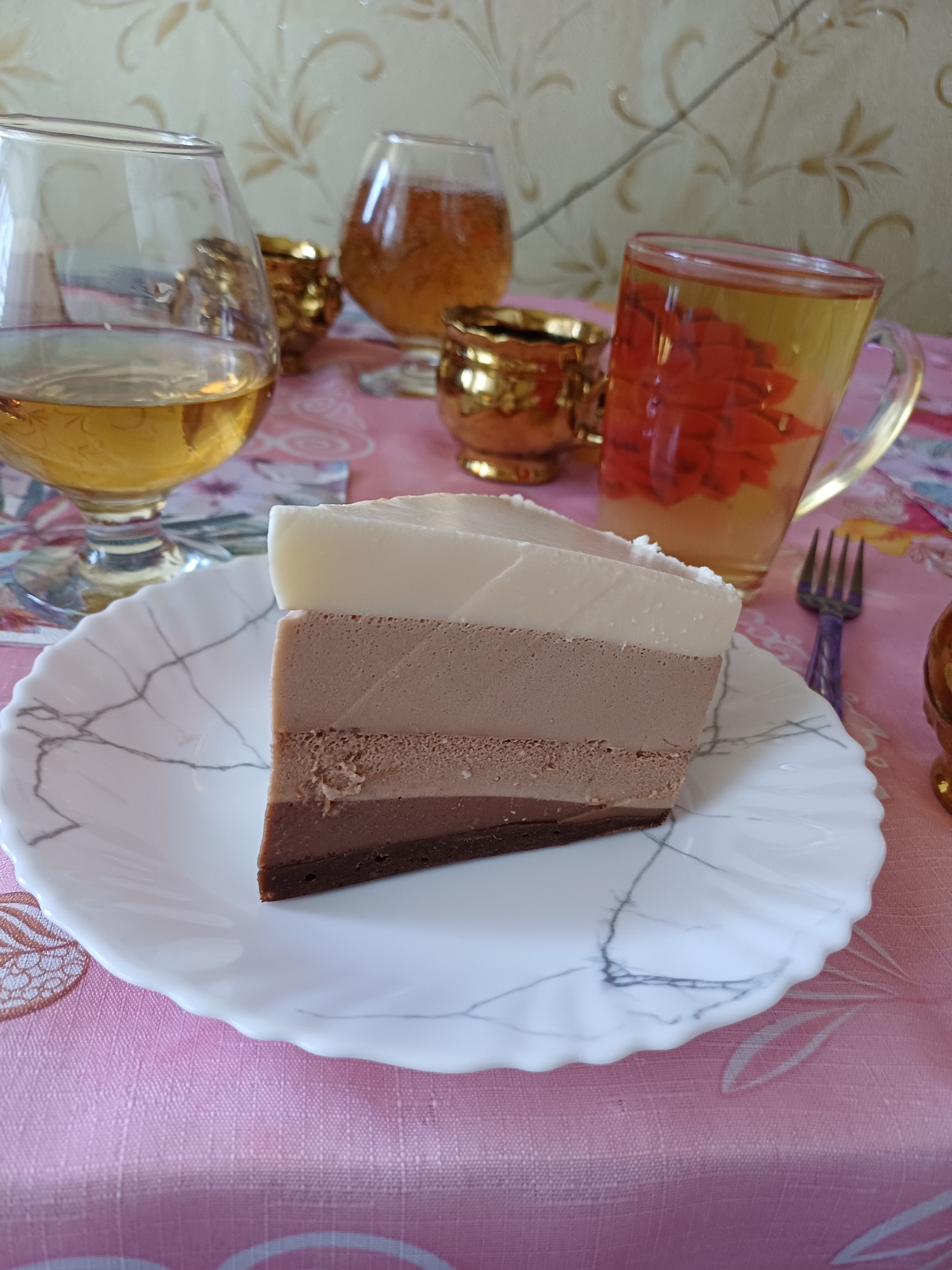 A slice of triple chocolate mousse cake rests on a white, scalloped plate with thin gray lines resembling marble. The cake is composed of three distinct layers: a pale white layer, a medium brown layer, and a dark brown layer. To the left of the plate are several glass objects with ornate gold bases, appearing to be cups or decorative items. One glass, in particular, is filled with an orange liquid that contains red, petal-like shapes. A purple fork with a pointed end is positioned above the cake and plate, partially obscured. The entire scene is set against a pink tablecloth with a subtle floral or leaf pattern. The lighting suggests it is daytime, likely indoors, given the reflection on the glassware. There is no visible text in the image. The overall impression is of a dessert presented for consumption or display, possibly in a home or a dining establishment.