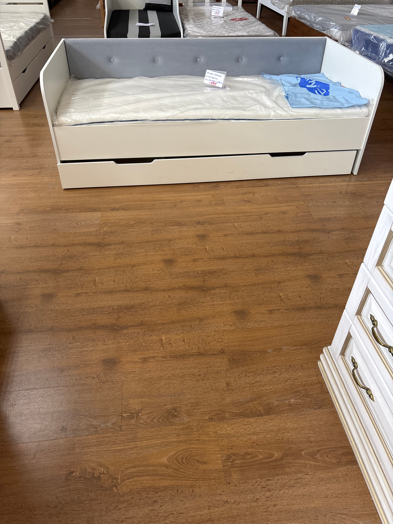 The image shows a white bed frame with a pull-out drawer. A mattress is inside the bed frame, wrapped in plastic. A blue cloth with a logo is draped over the mattress. The bed frame is positioned on a wooden floor. In the background, a white dresser with ornate handles is visible. The scene appears to be indoors, possibly in a furniture store or a bedroom. The lighting suggests daytime. There is text on a label attached to the blue cloth, which reads "детская коллекция" and some other Cyrillic characters. Another label visible on a mattress in the background has the number "5500" and Cyrillic text. The location context is Zaporizhzhia, Ukraine.