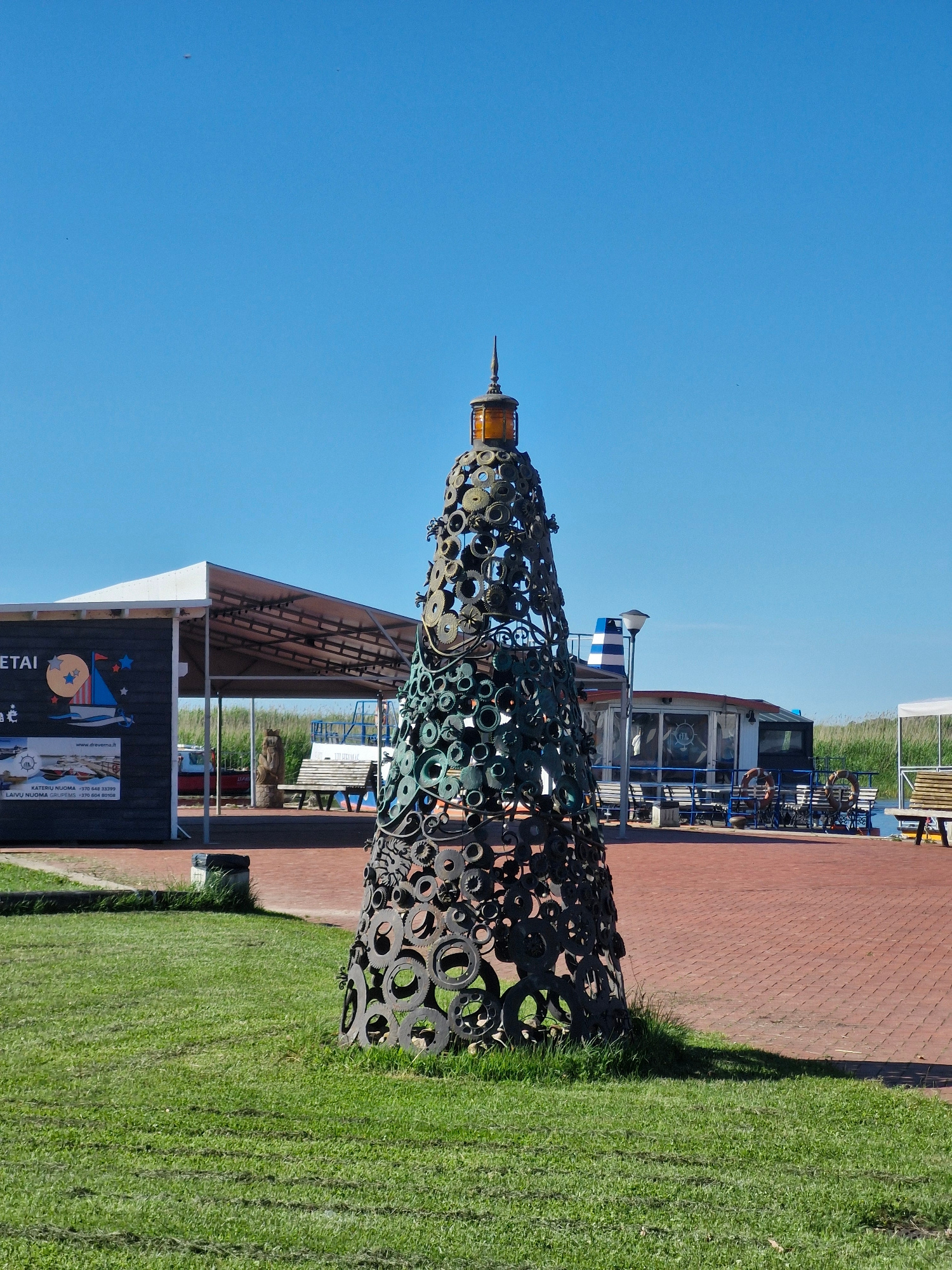 An abstract sculpture resembling a lighthouse, constructed from various metal components, stands in the foreground on a grassy area. The sculpture is dark and textured, with a prominent amber-colored element at its apex. Behind the sculpture, a paved brick walkway stretches towards a waterfront area. Along the walkway, there are buildings with awnings, outdoor seating, and boats docked in the water. A blue sky with sparse clouds dominates the upper portion of the image. The scene appears to be a public space, possibly a park or a marina, in Klišiai, Lithuania.