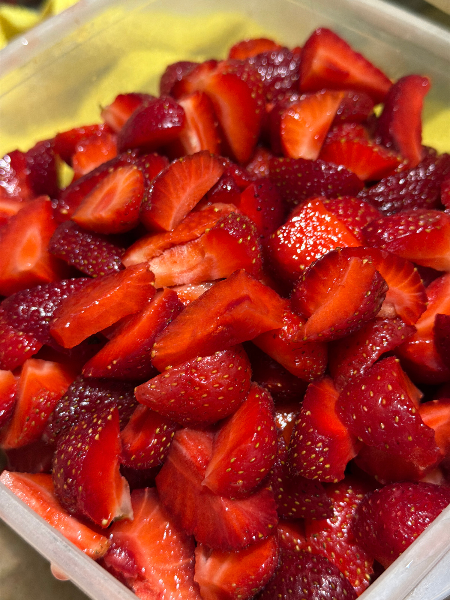 A close-up, high-angle, full-frame shot shows a clear plastic container filled with freshly sliced strawberries. The strawberries are a vibrant red, with some appearing to be whole halves and others cut into quarters. Their seeds are visible, and the cut surfaces reflect light, indicating moisture. The background is blurred and shows a bright yellow object, possibly fabric or a table. The lighting is overhead and somewhat diffused, highlighting the texture and glossiness of the fruit. The image captures a detailed view of the sliced strawberries within their container, with no people or other identifiable objects present.