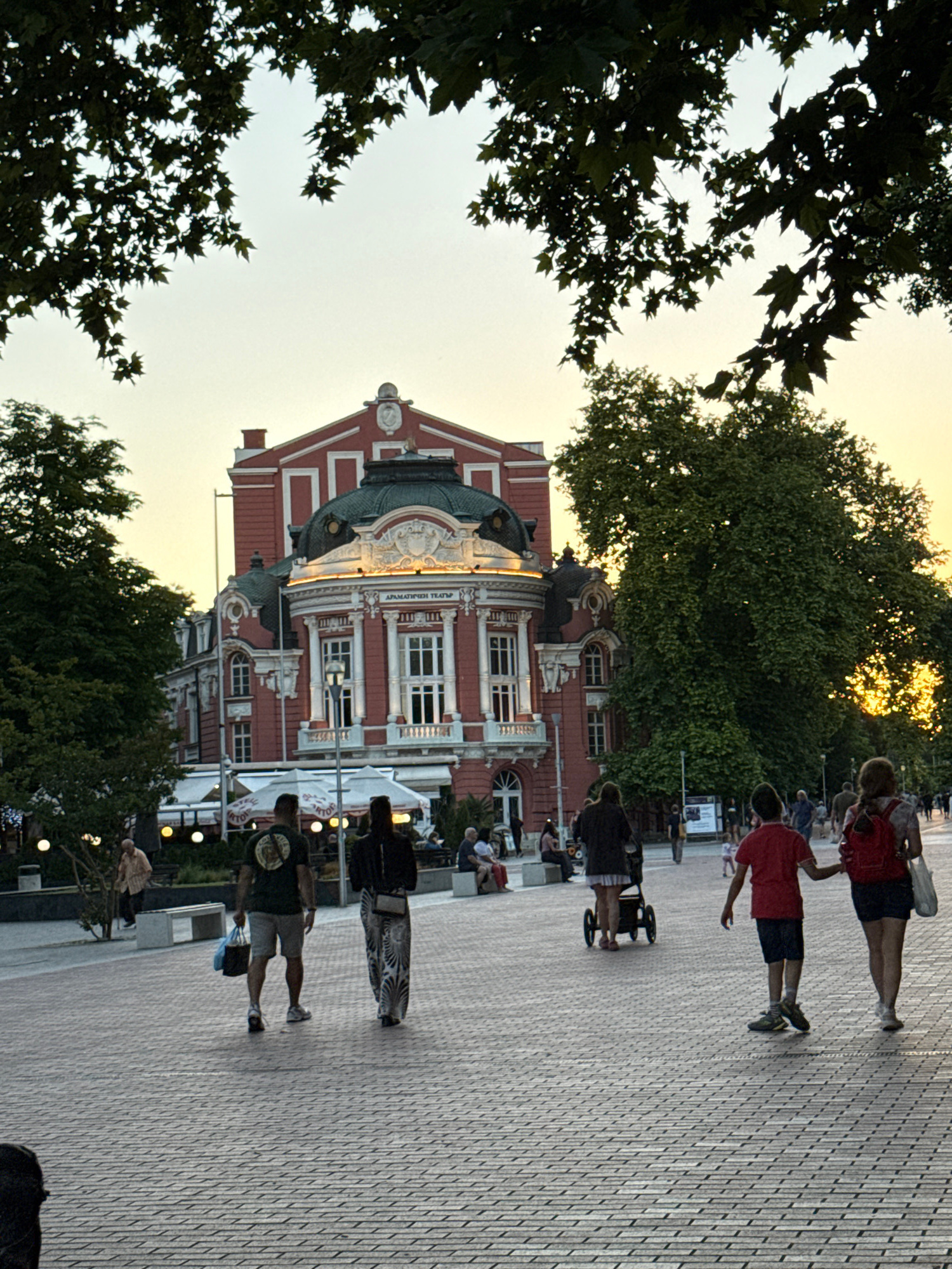 A paved pedestrian walkway with patterned brickwork is shown, leading towards a grand red building with a green dome and white architectural details. Trees with dark green leaves overhang the scene from above, framing the sky. Several people are walking on the walkway. In the foreground, a man and woman are walking away from the camera. The man is wearing a dark green t-shirt and grey shorts, carrying a blue bag. The woman is wearing a long, patterned dress. Further ahead, a woman is pushing a stroller. A young boy in a red t-shirt and dark shorts is walking with a woman wearing a red backpack. The building in the background appears to be a theater, with signage in a foreign language. Outdoor seating and umbrellas are visible in front of the building, suggesting a cafe or restaurant. The sky is a pale yellow, indicating either sunrise or sunset.