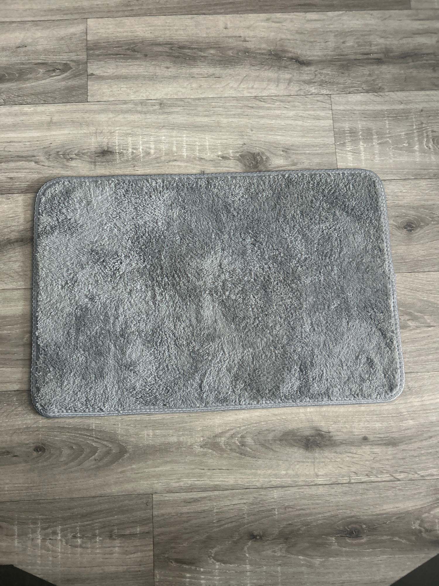 A rectangular gray bath mat with a textured, plush surface lies on a light gray wood-look laminate floor. The mat is positioned horizontally in the center of the frame. The flooring exhibits a grain pattern with variations in tone, creating a natural wood appearance. The image captures the mat from a top-down perspective. The scene is indoors and appears to be in a domestic setting. No people or specific landmarks are visible in the image.