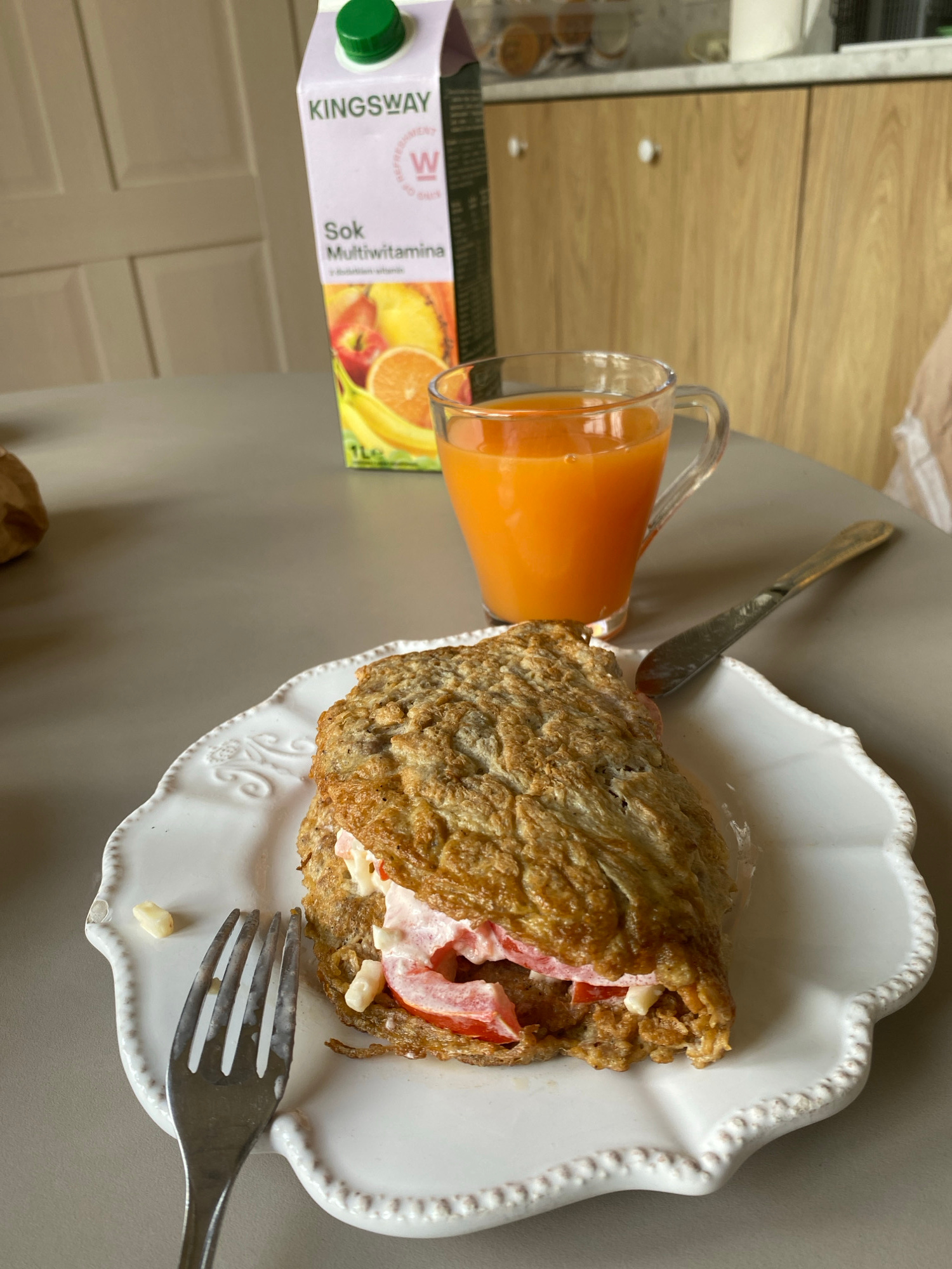 The image shows a breakfast setting on a grey surface, likely a table. The main subjects are a folded omelet filled with what appears to be tomato and a creamy sauce, served on a decorative white plate. To the left of the plate, a clear glass mug holds a vibrant orange juice. Next to the mug is a carton of "KINGSWAY Sok Multivitmina" juice, which is partially visible. A fork rests on the plate, with a small piece of food on one of its tines. A butter knife is placed on the table behind the mug. The lighting suggests it's daytime, possibly morning, and there are no obvious weather cues. The overall impression is that of a home-cooked meal, ready to be eaten. The visible text on the juice carton is "KINGSWAY", "Sok", and "Multivitmina". The setting appears to be indoors, possibly a kitchen or dining area, given the presence of cabinetry in the background that resembles kitchen units. The mood is casual and domestic.