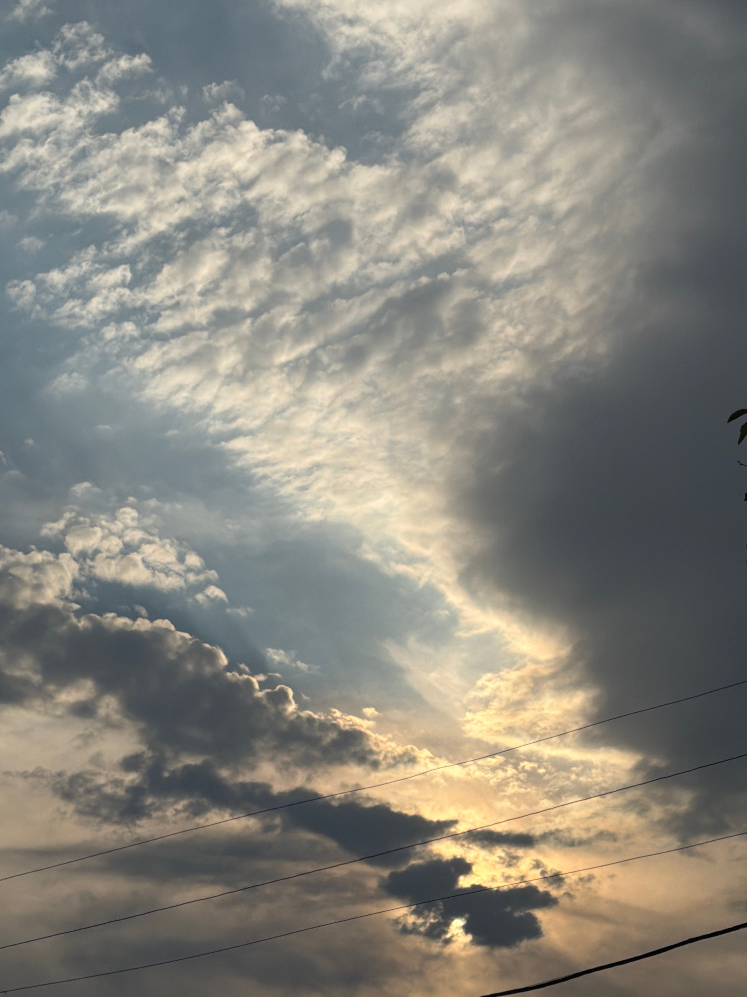 This image captures a dynamic sky with dramatic cloud formations, suggesting either the approach or departure of a storm. The sun is breaking through in the upper right portion of the frame, casting a warm, golden light on some of the clouds while others remain dark and ominous. Wispy, streaky clouds stretch across the left side of the image, contrasting with the heavier, more dense formations on the right. Several power lines are visible, stretching diagonally across the frame, indicating an outdoor, likely urban or semi-urban, setting. The overall mood is one of awe and perhaps a hint of foreboding, typical of dramatic weather phenomena. The time of day appears to be either sunrise or sunset, given the low angle of the sun and the quality of light. There is no visible text in the image. The location context of Tashkent, Uzbekistan, does not provide specific visual cues within the image itself but can inform the general atmospheric conditions that might be expected.