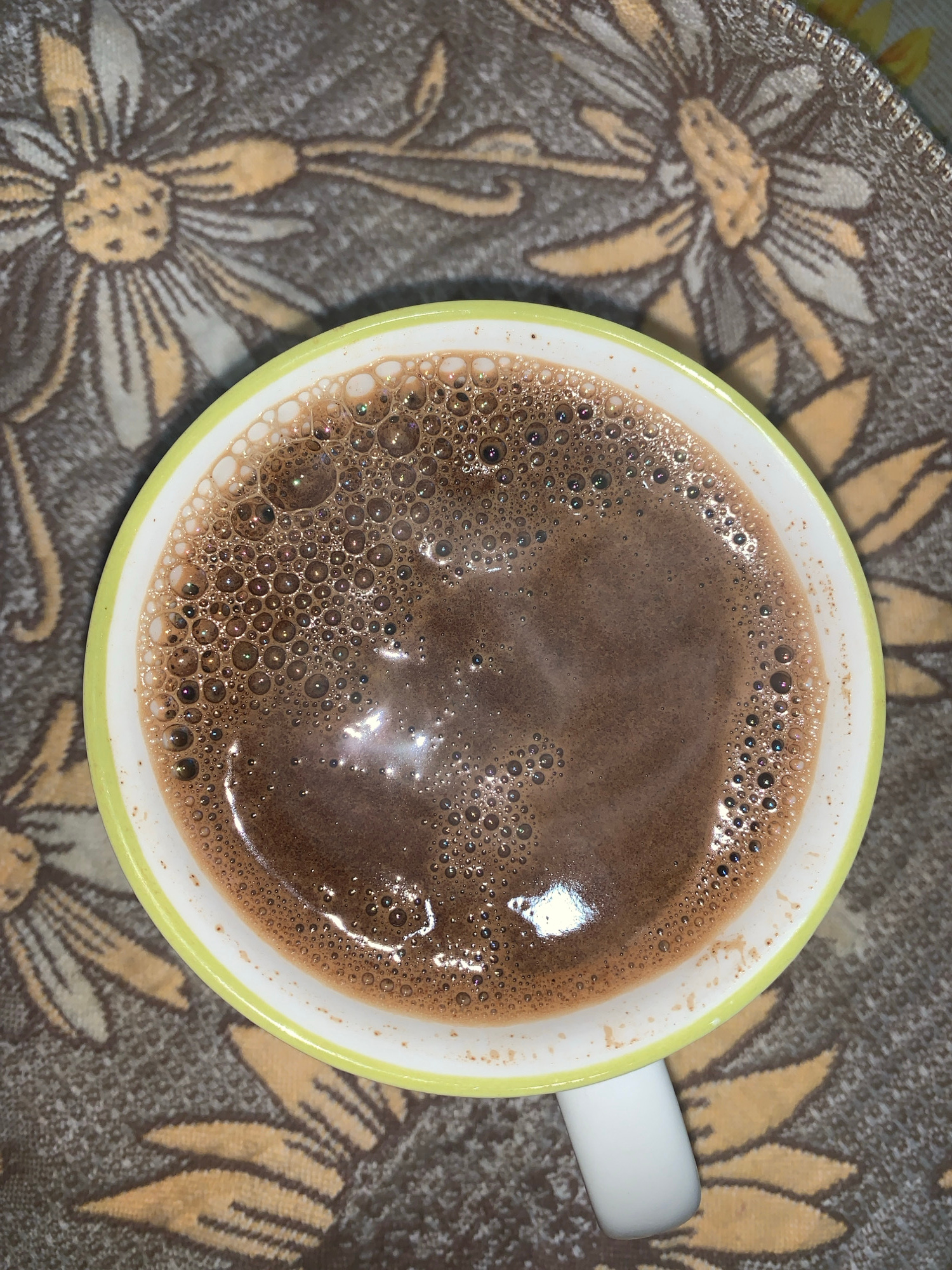 A close-up, overhead shot shows a mug of hot chocolate with a foamy surface. The mug has a white interior and a lime-green rim. The mug rests on a patterned fabric, which appears to be a tablecloth or placemat, featuring stylized yellow and brown flowers. The fabric has a dark brown background. The hot chocolate is a rich brown color with a multitude of small bubbles on its surface, some reflecting light. The image is focused on the mug and its contents, with the patterned fabric slightly blurred in the background. The scene is indoors, likely on a table.
