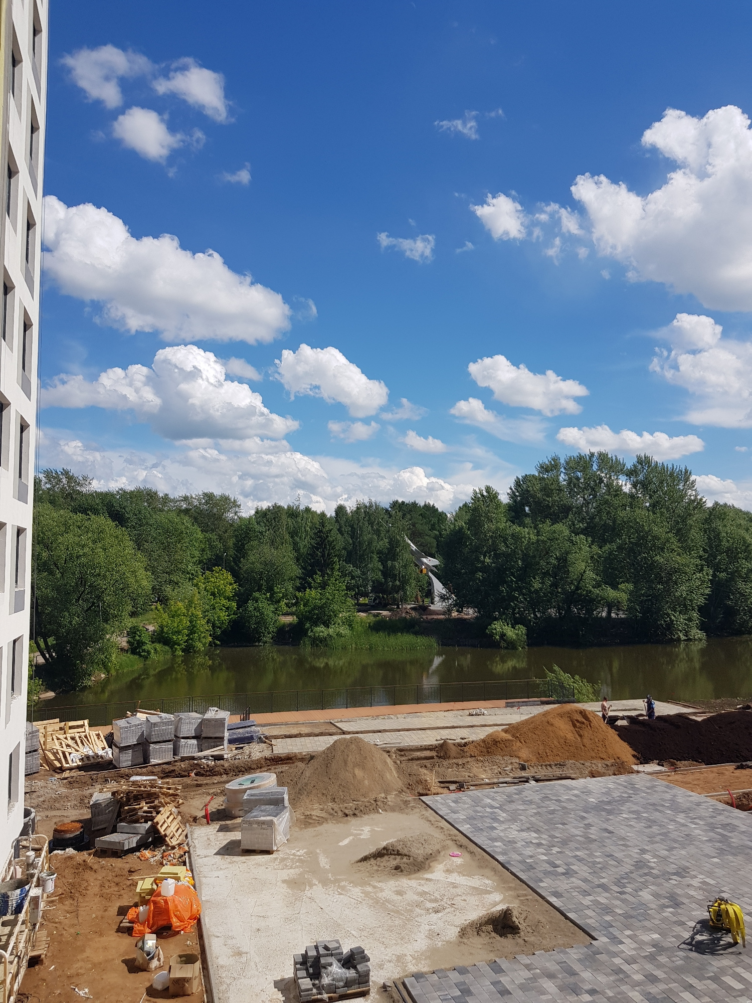 A vertical, eye-level, medium shot captures a construction site adjacent to a body of water with lush green trees and a bright blue sky filled with white, fluffy clouds. In the foreground to the left, the side of a white building with dark-framed windows is visible. The construction area is littered with materials, including stacks of gray pavers wrapped in plastic, wooden pallets, piles of sand and dirt, and orange tarps. A yellow compaction machine sits on a paved area to the right of the frame. In the middle ground, a river or lake is bordered by a dark fence. Beyond the water, a dense forest of green trees stretches across the horizon, with a white structure resembling an airplane or a monument visible amongst the trees on the left. Two figures, one in a dark blue shirt and another in a light-colored shirt, are standing near a pile of dirt on the right side of the river bank.