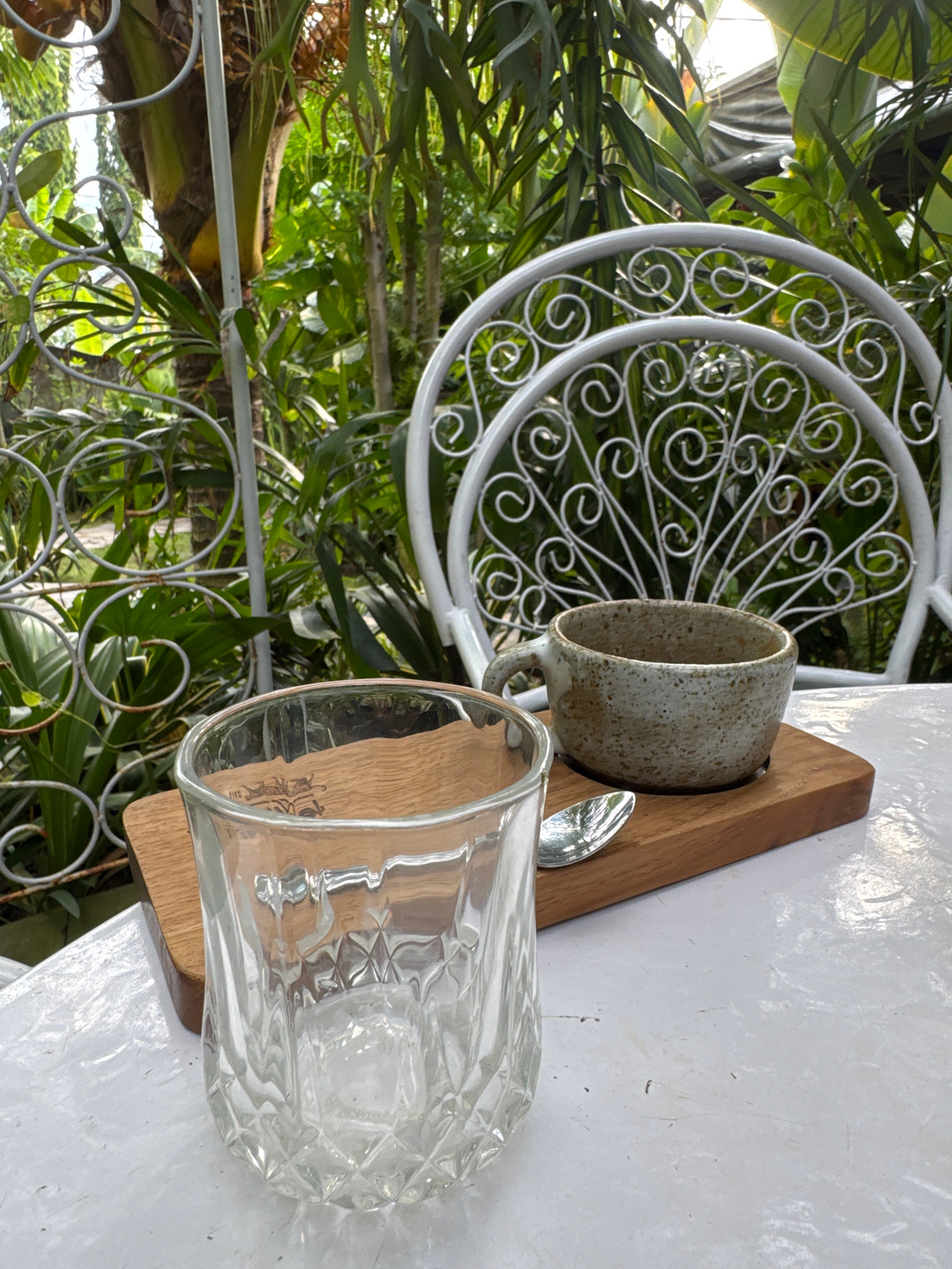 A close-up, eye-level shot captures a rustic coffee setting on a white table, set against a lush, green backdrop of tropical foliage and a decorative white metal chair. In the foreground, a clear, textured glass tumbler sits empty on a small wooden board. To its right, on the same board, is a small, speckled ceramic cup with a handle, presumably containing coffee. A silver spoon rests beside the cup. The background reveals a vibrant garden with palm trees and other plants, with a glimpse of a white metal trellis or fence. The lighting suggests it is daytime. The scene appears to be an outdoor cafe or a serene garden patio in Abang, Indonesia, possibly a local eatery or a private residence with a garden view. No people are visible in the image.