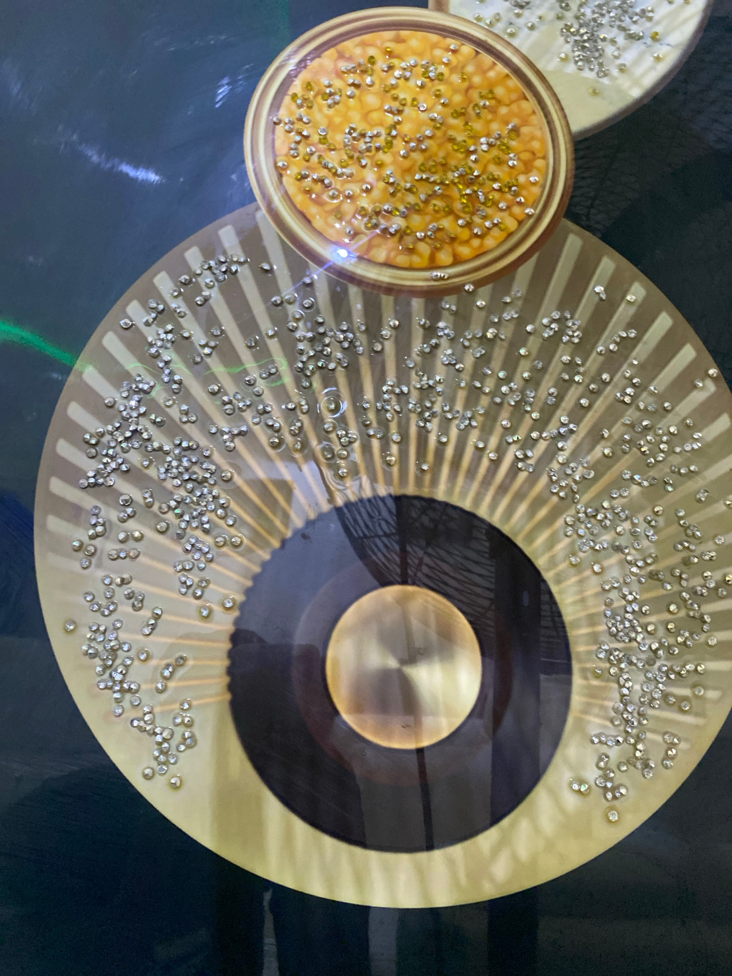 The image depicts a close-up, overhead view of two decorative, circular objects against a dark, reflective background. The primary object is a large, golden fan-like structure adorned with numerous small, clear, faceted embellishments, resembling rhinestones or small crystals. These embellishments are scattered unevenly across the fan's surface, concentrated more densely towards the upper portion. The fan has a ribbed design radiating from its center.
Nestled on top of this fan is a smaller, round object with a raised rim, containing a textured, yellowish-orange substance. This substance also appears to be decorated with similar clear and yellowish faceted embellishments, some of which have a more pronounced yellow hue. The smaller object has a golden rim.
The background is dark and highly reflective, showing subtle streaks of light, possibly from artificial illumination. There are also faint green light streaks visible in the upper left portion of the image. The overall impression is one of decorative craftsmanship, possibly an art installation or a piece of interior design.
There are no people, animals, or explicit indications of a specific city or location within the image itself. The details provided in the prompt about "Giginyu, Nigeria" are not visually supported by the content of the photograph.