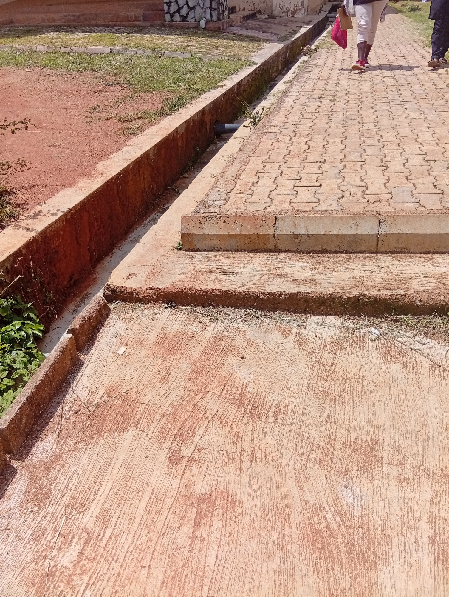 The image captures an outdoor scene in Gwong, Nigeria, featuring two individuals walking on a paved walkway. The walkway is constructed from interlocking brick pavers in a tan color, arranged in a repeating pattern. To the left of the walkway, there is a concrete drainage channel with reddish-brown earth and some greenery growing along its edges. Beyond the drainage channel, a dirt area with sparse grass is visible, leading towards a building with stepped brick foundations. The building's facade appears to be decorated with light-colored stones.
The two people are seen from the waist down, walking away from the viewer. The person in front is wearing white trousers and red sneakers, carrying a red bag. The person behind them is wearing dark trousers and sandals. The lighting suggests it is daytime, with shadows present, indicating a sunny or partly cloudy day. The overall impression is of a functional outdoor space, possibly a public area or pathway on a campus. There is no visible text in the image.
