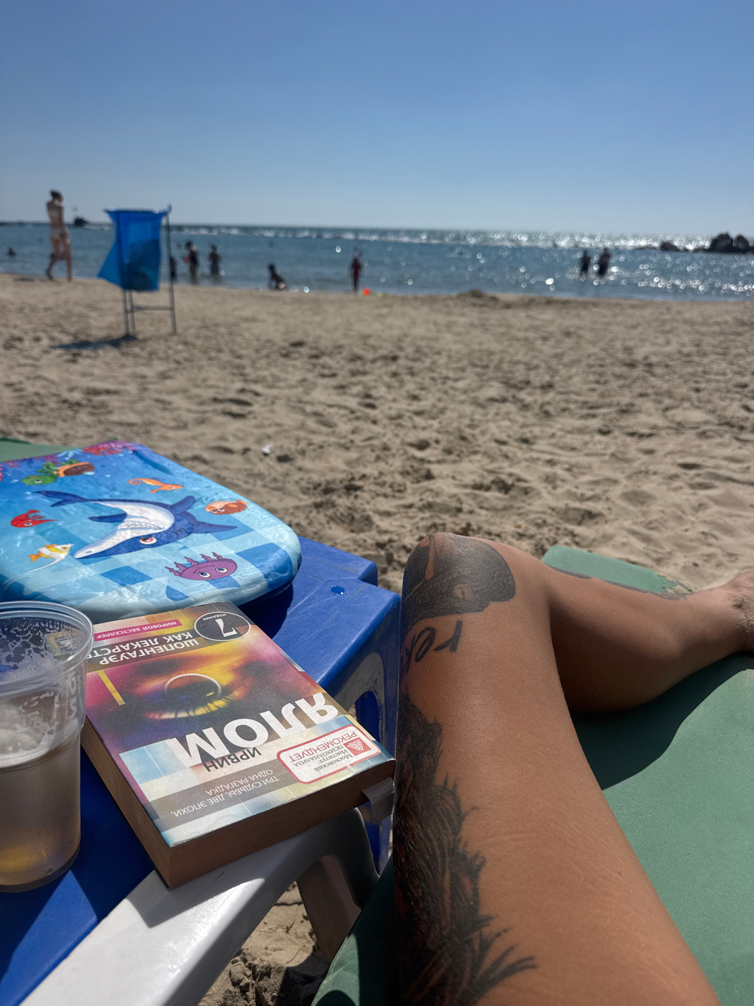 The image shows a close-up of a person's leg with tattoos, resting on a beach chair. Next to the leg is a book and a glass of beer. The beach is visible in the background, with people swimming in the ocean and a blue trash can on the sand. The sky is clear and blue. The scene appears to be a relaxing day at the beach.