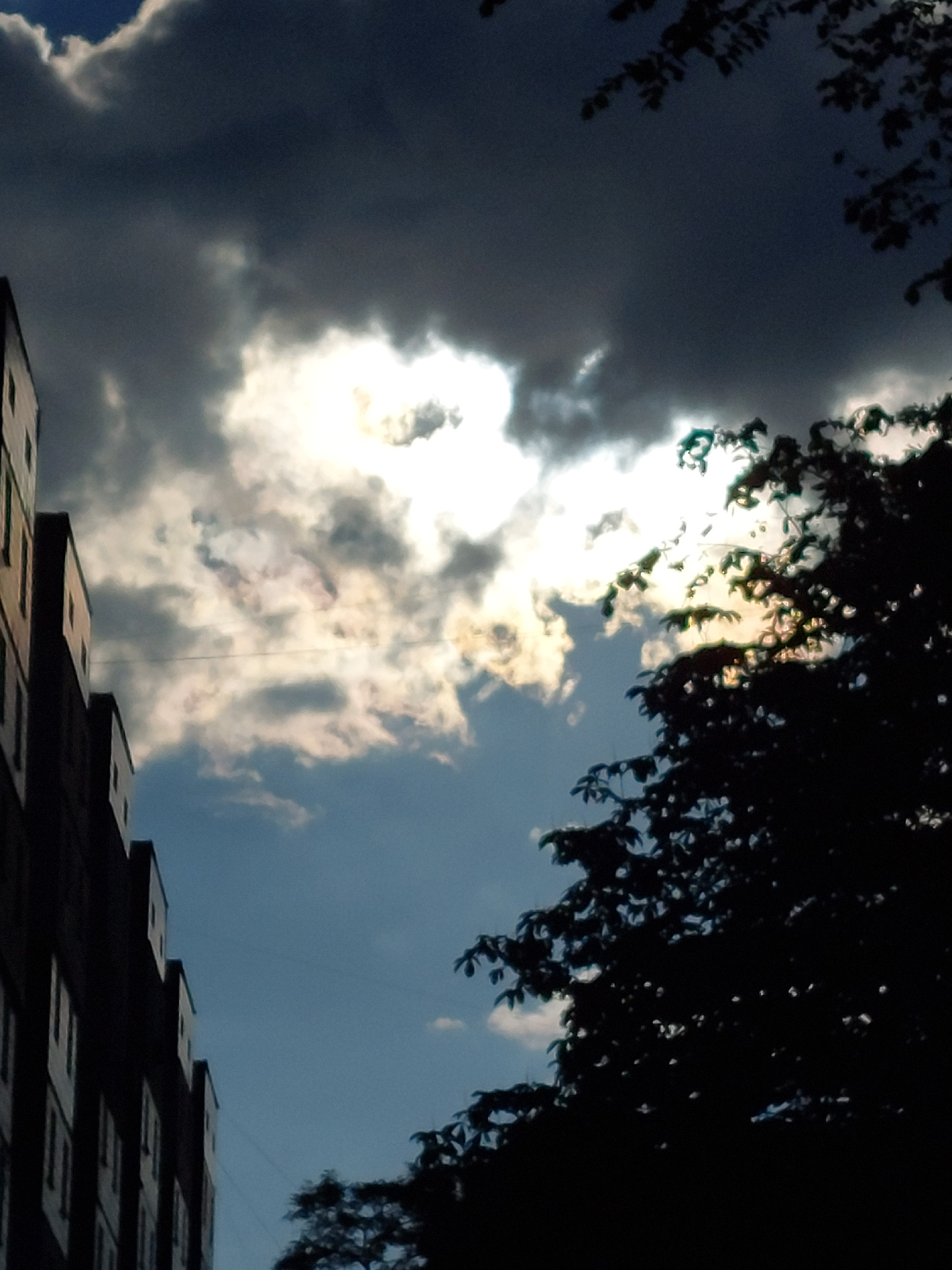 A low-angle, vertical shot captures a dramatic sky over a city. Dark, heavy clouds dominate the upper portion of the frame, with a bright patch of sunlight breaking through in the center, casting an ethereal glow. To the left, the dark silhouette of a multi-story building with visible windows extends upwards. On the right, the dense, dark foliage of a tree partially obscures the sky, creating a stark contrast with the illuminated clouds. The lower portion of the image reveals a lighter blue sky. The scene appears to be outdoors in an urban environment.