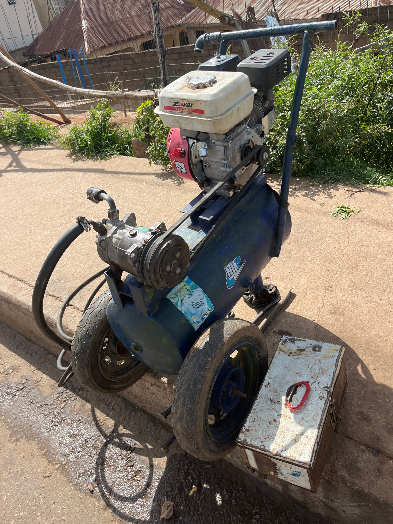 An outdoor, eye-level, medium shot captures a portable air compressor parked on a dirty roadside in Bukuru, Nigeria. The compressor, primarily blue with a white gas tank and red starter, features a black belt connecting its engine to the compressor pump. A black hose extends from the pump, and two weathered tires support the unit. To the right of the compressor, a small, white metal toolbox with a red carabiner clip rests on the ground. The background reveals a brick wall, a corrugated metal roof, and dense green vegetation, suggesting a rural or semi-urban environment. The ground is a mixture of dirt and pavement, with shadows cast by the compressor and the midday sun.
