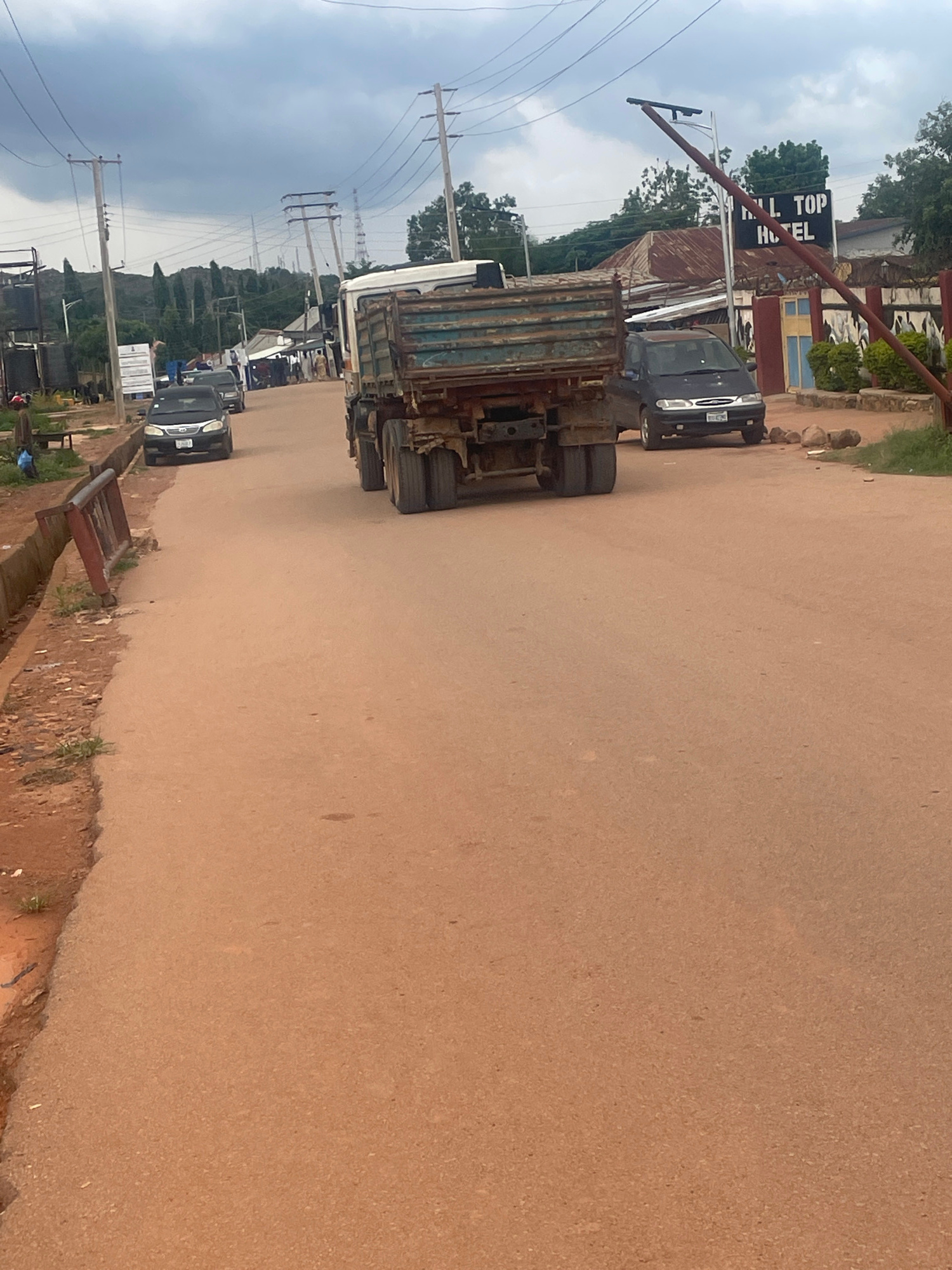 The image depicts a street scene in Gura Top, Nigeria, with several vehicles present. Dominating the foreground is the back of a rusty, green truck with a visible trailer. To the left of the truck, a dark-colored car is parked. Further down the road, a line of other vehicles is visible, including a black car in the middle ground.
In the background, there are buildings, including one with a sign that reads "HILL TOP HOTEL." Trees and foliage are scattered throughout the scene, and power lines stretch across the sky, suggesting an urban or semi-urban environment. The sky is overcast, indicating cloudy weather. The road surface appears to be made of dirt or gravel, typical of many roads in Nigeria.
The image is taken from a slightly elevated perspective, looking down the road. No people are clearly visible in the image, although the presence of parked cars suggests activity in the area. The overall impression is a typical roadside scene in a Nigerian town, with a mix of commercial and residential structures.