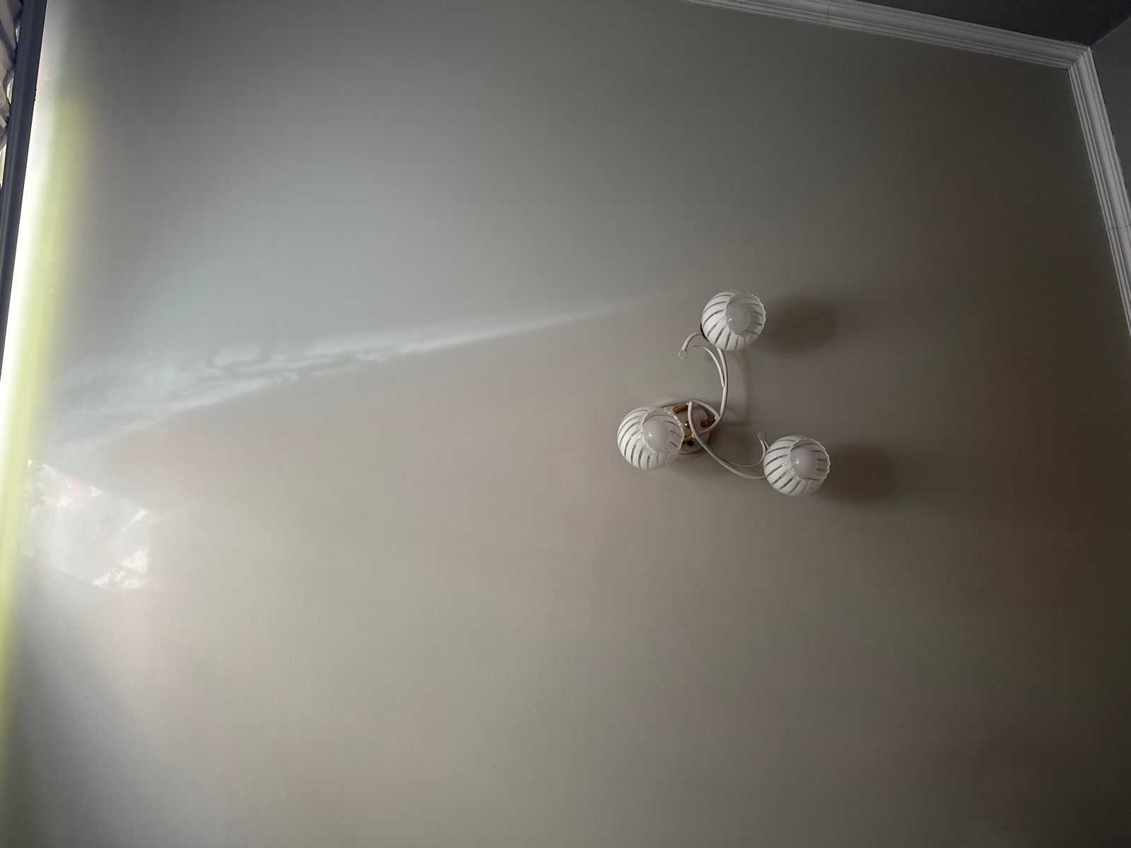 A white, three-headed ceiling light fixture is mounted on a light beige wall. The light fixture has a curved, branching design, with each head featuring a white, spherical lampshade with a slatted texture. A beam of light is casting a diagonal pattern across the wall from the left side of the image, likely from a window or another light source. The ceiling is visible at the top of the image, with white crown molding separating it from the wall. The room appears to be indoors.
