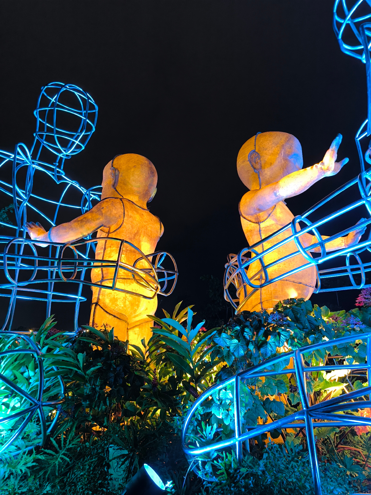 This image shows a nighttime outdoor installation featuring three illuminated figures. The central figure is a large, bright yellow sculpture of a baby, facing to the right and reaching out its left arm. To the left of the baby is another, taller, wireframe sculpture of an adult figure, made of blue neon lights, with its arms reaching towards the baby figure. To the right of the baby is a second, identical yellow baby sculpture, facing left and reaching out its right arm. All three figures are partially enclosed by blue wireframe structures. The sculptures are surrounded by lush green foliage, illuminated by blue and yellow lights. The background is dark, suggesting a night sky. The installation is located in the city of Kediri, Indonesia, specifically at the Gor Jayabaya public facility.