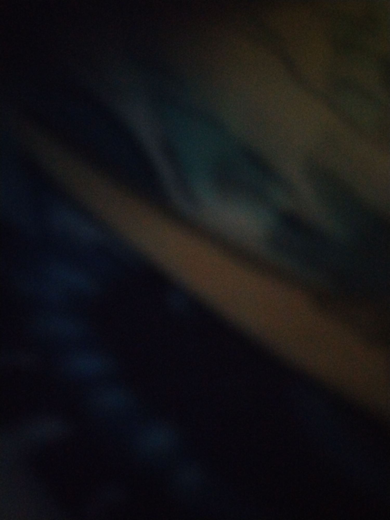 The image is a close-up, abstract shot with a shallow depth of field, showcasing soft focus and blended colors. The dominant hues are dark blues, blacks, and muted browns, with diagonal streaks of lighter color suggesting some form of illumination or material texture. The lighting is dim and diffused, creating a moody and atmospheric effect. There are no discernible objects, people, or actions depicted. The scene appears to be a textured surface, possibly fabric or a painted wall, rendered abstract by the photographic technique. The location is unknown.