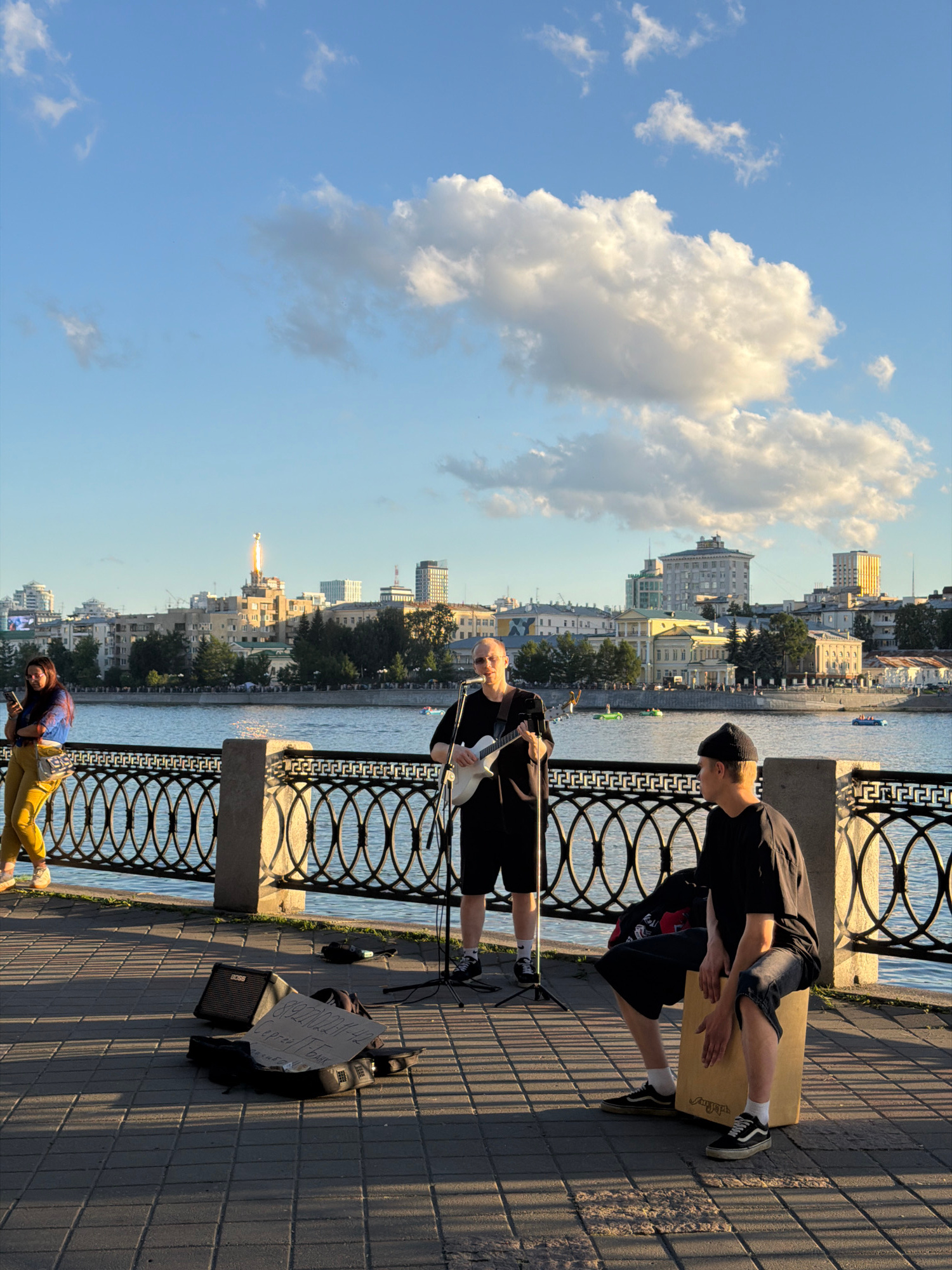 This is a medium shot photograph taken from a slightly elevated perspective, capturing a street performance scene on a riverside promenade in Yekaterinburg, Russia. The sun is low in the sky, casting long shadows and illuminating the scene with a warm golden light.
In the foreground, a paved walkway made of square bricks is visible. On the walkway, three individuals are present. The person on the left is partially visible from the waist down, wearing yellow pants and a blue and black patterned shirt, holding a phone and leaning against a decorative black metal railing. To their right, a person stands playing a white electric guitar and singing into a microphone. They are wearing a black t-shirt and shorts. In front of them, on the ground, are a black guitar case, an amplifier labeled "BOSS," and a sign with Cyrillic text that appears to be contact information or a donation request. To the right of the guitarist, another person is seated on a cajon drum, wearing a black t-shirt and dark shorts, and a black beanie. They are looking to the right, away from the camera.
Behind the performers and the railing, a wide body of water, likely the Iset River, stretches across the middle ground. Several small boats are visible on the water. Across the river, the Yekaterinburg skyline is prominent, featuring a mix of architectural styles. Notable buildings include a taller structure with a distinctive spire and what appears to be a light source emanating from its top, resembling a beacon or light show. Other buildings vary in height and design, with some exhibiting classical architectural elements and others being more modern. Trees are scattered throughout the city landscape.
The sky above is a clear blue with scattered white and gray clouds, some of which are illuminated by the setting sun. The overall atmosphere is peaceful and vibrant, suggesting a typical evening in a city park or along a waterfront area.