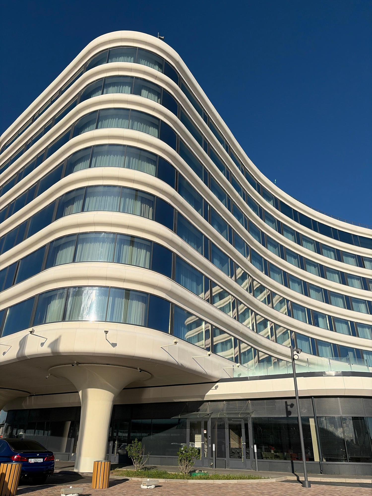 This media file captures a modern architectural structure with a distinctive curved facade, bathed in sunlight against a clear blue sky. The building's exterior features a repeating pattern of light-colored vertical supports and large, reflective windows, creating a sense of rhythmic movement and depth. The curved design gives the impression of a wave or a sail, emphasizing a contemporary aesthetic.
The scene is set outdoors, with the bright sunlight suggesting a clear day. Shadows are cast on the building's facade and the ground, indicating it's likely daytime. The ground level shows a paved area with some greenery and outdoor seating elements, hinting at a public space or commercial area. A dark blue car is visible parked on the right side of the frame, with a license plate that appears to be Russian, displaying Cyrillic script and numbers "050 PRG".
The overall mood is one of modern sophistication and architectural grandeur. The clean lines and reflective surfaces create a bright and airy atmosphere. There are no people visible in the frame, focusing the attention solely on the building and its surroundings. The architectural style and the presence of Cyrillic script on the license plate align with the provided location context of Saint Petersburg, Russia. The building's design appears to be the primary subject, showcasing innovative urban architecture.