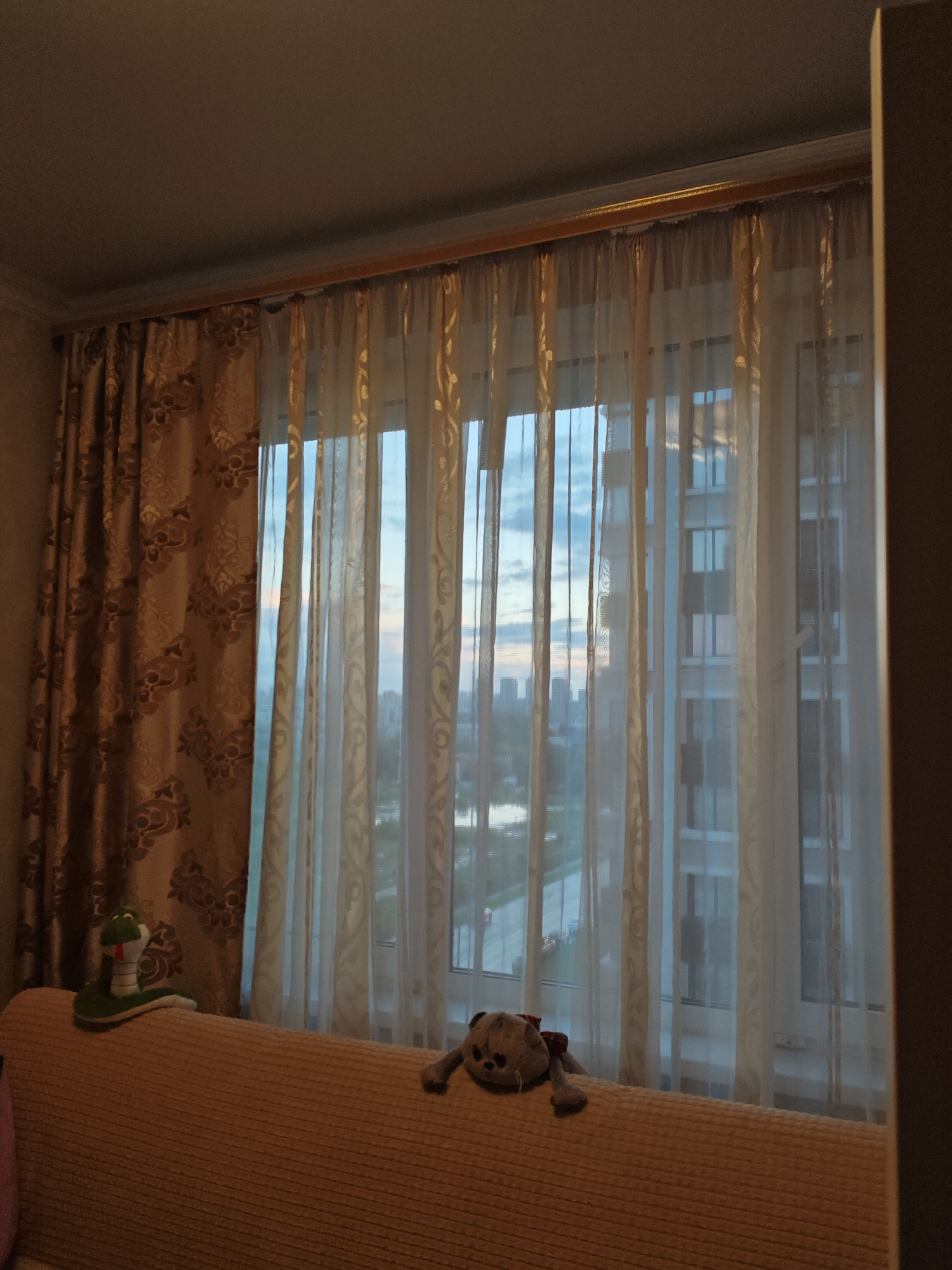 The image shows a view from inside a room looking out a window covered by two layers of curtains. The outer curtains are a heavy, patterned fabric in shades of gold and brown. The inner curtains are sheer and white, allowing some light to pass through. Outside the window, a cityscape is visible with tall buildings and a cloudy sky. In the foreground, the top of a light brown couch is visible, with a stuffed snake toy on the left and a stuffed bear toy on the right. The lighting suggests it may be dusk or early morning.