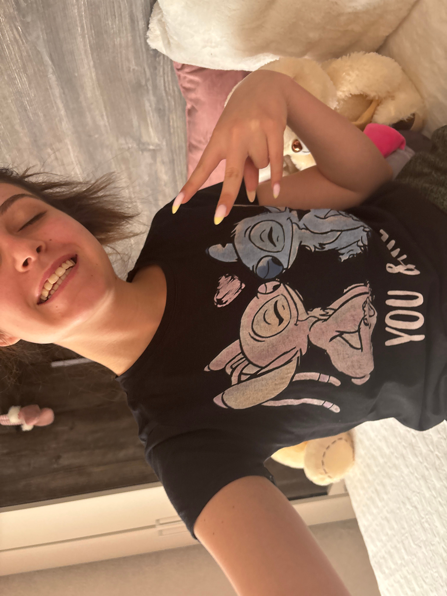 A woman is taking a selfie in a bedroom. She is lying on a bed with her head towards the bottom of the frame, and the camera is angled slightly downwards. She is wearing a black t-shirt with a graphic of Stitch and Angel from Lilo & Stitch on it, along with the text "YOU & I". The woman has long, dark hair, and is smiling with her eyes closed. Her nails are long and painted with a French manicure.
The bed is made with a white duvet. To the left of the woman, there are several stuffed animals, including a large white one and a smaller yellow one with pink ears. A pink pillow is also visible. The flooring in the room appears to be dark wood.
There are no specific time of day or weather cues visible. The general mood is relaxed and playful, suggested by the smile and the presence of the stuffed animals. The location context of Bulgarovo, Bulgaria, is provided.