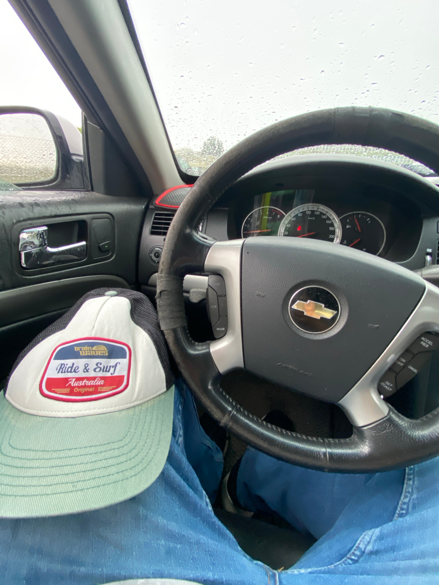 The image shows a first-person perspective from inside a car, with a focus on the steering wheel and a cap. The steering wheel is black leather with a silver Chevrolet emblem in the center. The dashboard displays gauges, including a speedometer, tachometer, and fuel gauge, with the needle on the speedometer indicating a speed of approximately 80 km/h. The car's interior appears to be in normal condition, with some visible wear on the steering wheel.
A white and black trucker cap with a light green brim rests on the driver's lap. The cap features a red and white patch with text that reads "brain waves," "Ride & Surf," "Australia," and "Original."
The scene is set on a rainy day, as evidenced by the water droplets on the windshield and the side mirror. The environment outside the car appears to be a street or road, with some blurred greenery visible.
This image does not provide any information to determine the specific location in the city of Police, Poland. The details visible are generic to a car interior and a rainy day setting.
