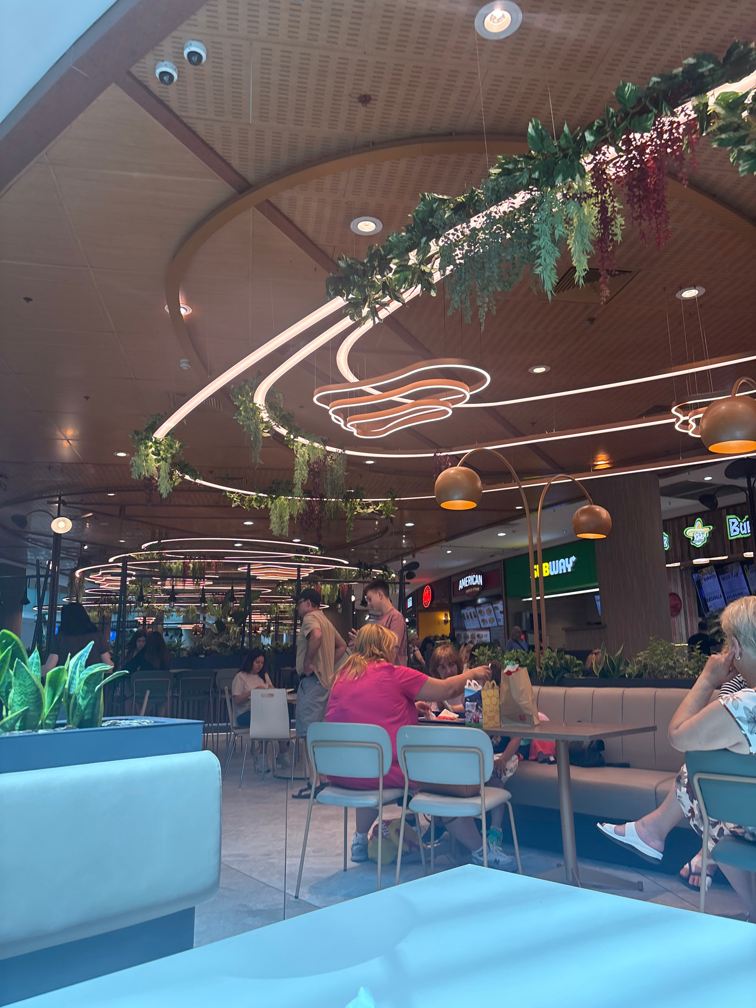 The image captures a brightly lit indoor dining area, likely a food court or a restaurant with an open-concept design. The ceiling features a distinctive wooden paneling and is adorned with hanging artificial greenery, creating a modern, biophilic aesthetic. Circular neon light fixtures and large pendant lamps with golden shades add to the contemporary ambiance.
In the foreground, a light blue bench or seating area is visible. Several people are seated at tables further back. A group of individuals, including two women in pink and floral tops, are seated at a table, engaged in conversation and eating. Another woman in a floral dress is seated on the right side of the frame. In the background, food establishments like "Subway" and "American" are identifiable by their signage. The overall environment suggests a bustling and well-maintained public space designed for dining and socializing. The lighting is bright, contributing to a lively and welcoming atmosphere.