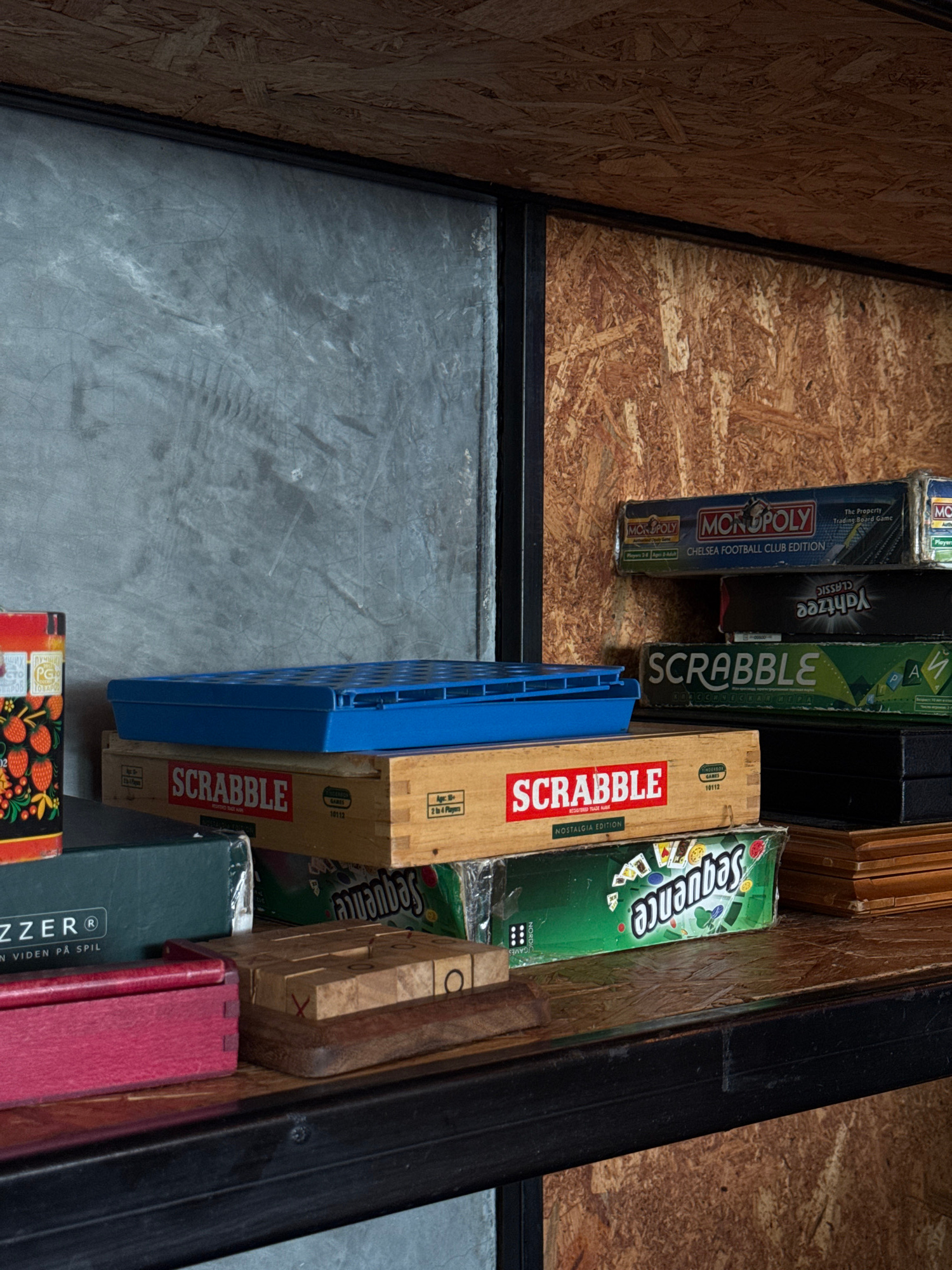 This is a close-up, high-angle shot of several board game boxes stored in a somewhat cluttered environment. The games are stacked vertically on a wooden-slatted shelf or surface. Visible game titles include "MONOPOLY" (specifically the "Chelsea Football Club Edition"), "Yahtzee Classic," "SCRABBLE" (in both a standard and "Nostalgia Edition"), and "Sequence." There is also a blue plastic game piece or tray positioned in front of some of the boxes. The background is a textured wooden surface, likely particleboard or plywood. The floor in the foreground appears to be concrete. The lighting is even and does not suggest a specific time of day. The overall impression is one of storage or a collection of games.
