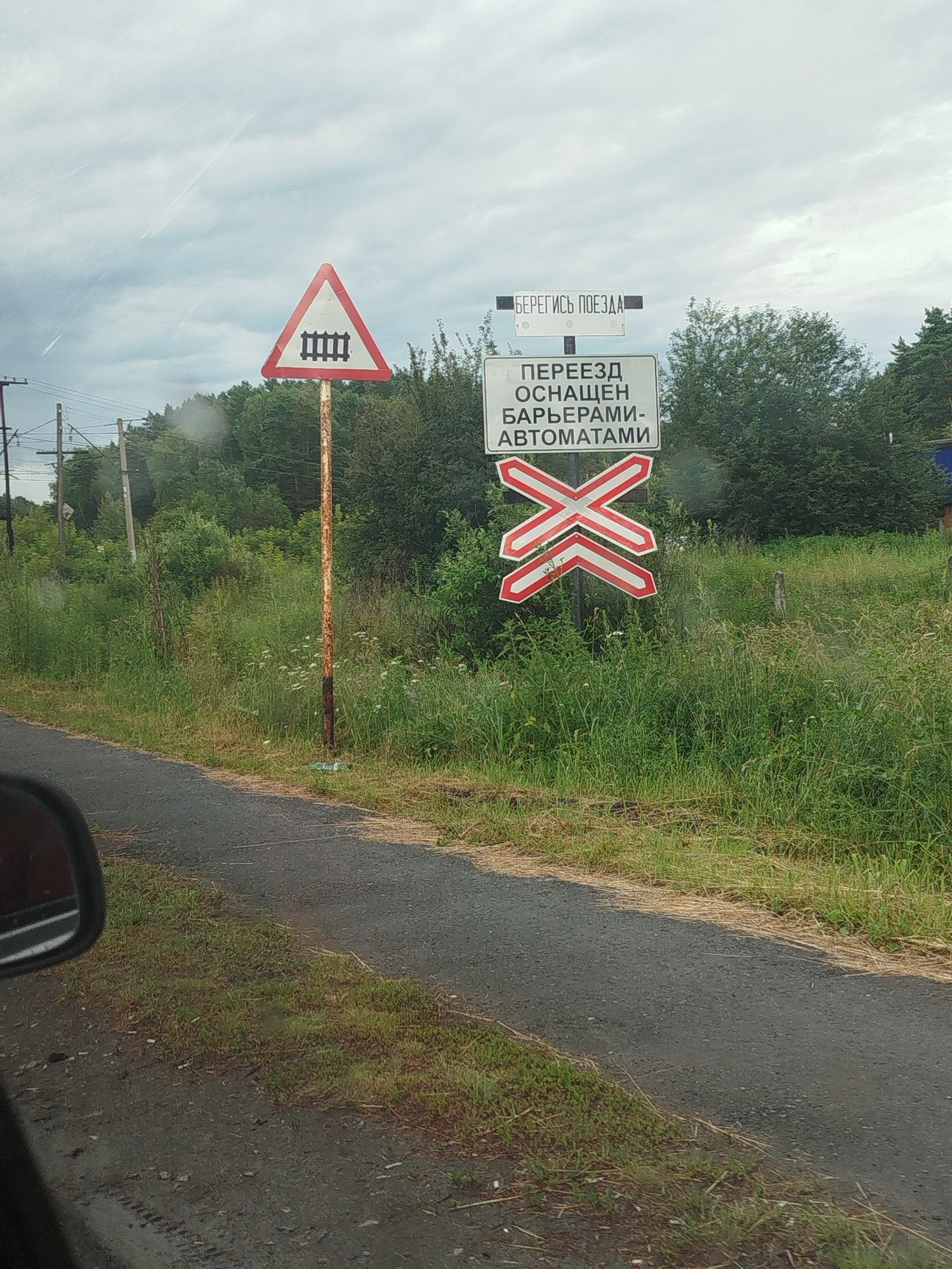 A view from inside a vehicle shows a rural road with a railroad crossing and associated signage. The road is paved and wet, indicating recent rain or damp conditions. To the left of the road, tall green grass and vegetation are visible. On the right side of the road, several signs are posted on a rusty metal pole. The topmost sign is a red and white triangle with a black depiction of train tracks. Below this is a rectangular white sign with black text that reads "БЕРЕГИСЬ ПОЕЗДА" (Beware of the train). Further down is another rectangular white sign with black text which reads "ПЕРЕЕЗД ОСНАЩЕН БАРЬЕРАМИ-АВТОМАТАМИ" (The crossing is equipped with automatic barriers). Below this are two red and white striped "X" shaped signs, indicating a single railroad track. Behind the signs, a dense line of trees forms the background. In the far distance, power lines are visible. The sky is overcast and grey. The scene appears to be located on the outskirts of Zavodoukovsk, Russia.