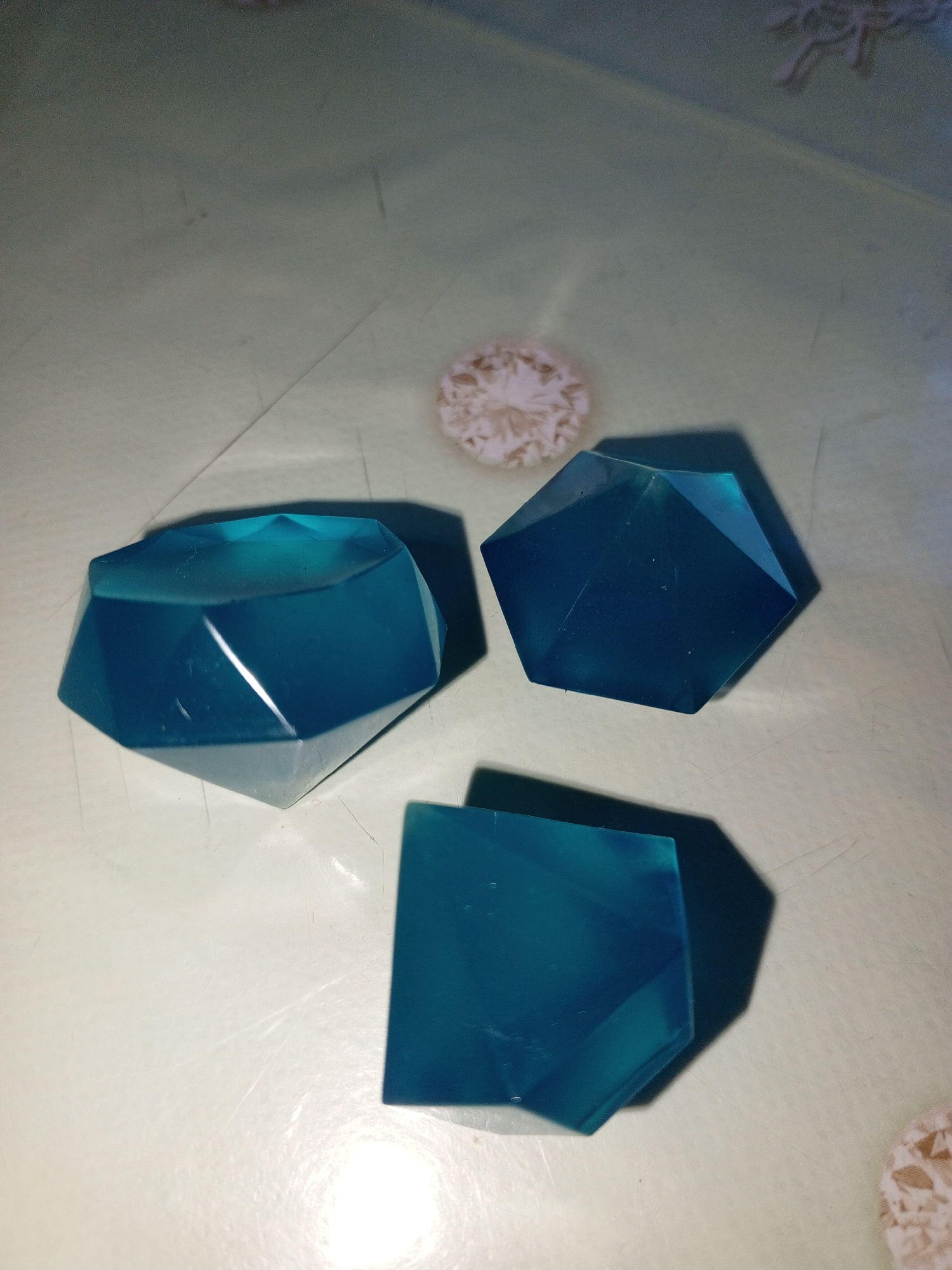 The image shows three geometric, faceted, translucent blue objects resembling gemstones. They are arranged on a light-colored, textured surface, possibly a tablecloth or placemat. One object is in the foreground, appearing as a square or rectangular prism with beveled edges. To the left and slightly behind it is a larger, faceted object with a rounded top. To the right and further back is another faceted object, similar in shape to the one on the left. In the upper center of the image, there is a small, light-colored, circular object with a delicate, floral pattern. The lighting suggests it is daytime, with shadows cast by the objects indicating a light source from the top right. The overall scene is still and decorative, with no clear activity taking place. There is no visible text in the image. The location context of Saint Petersburg, Russia, does not provide specific visual cues within the image itself.