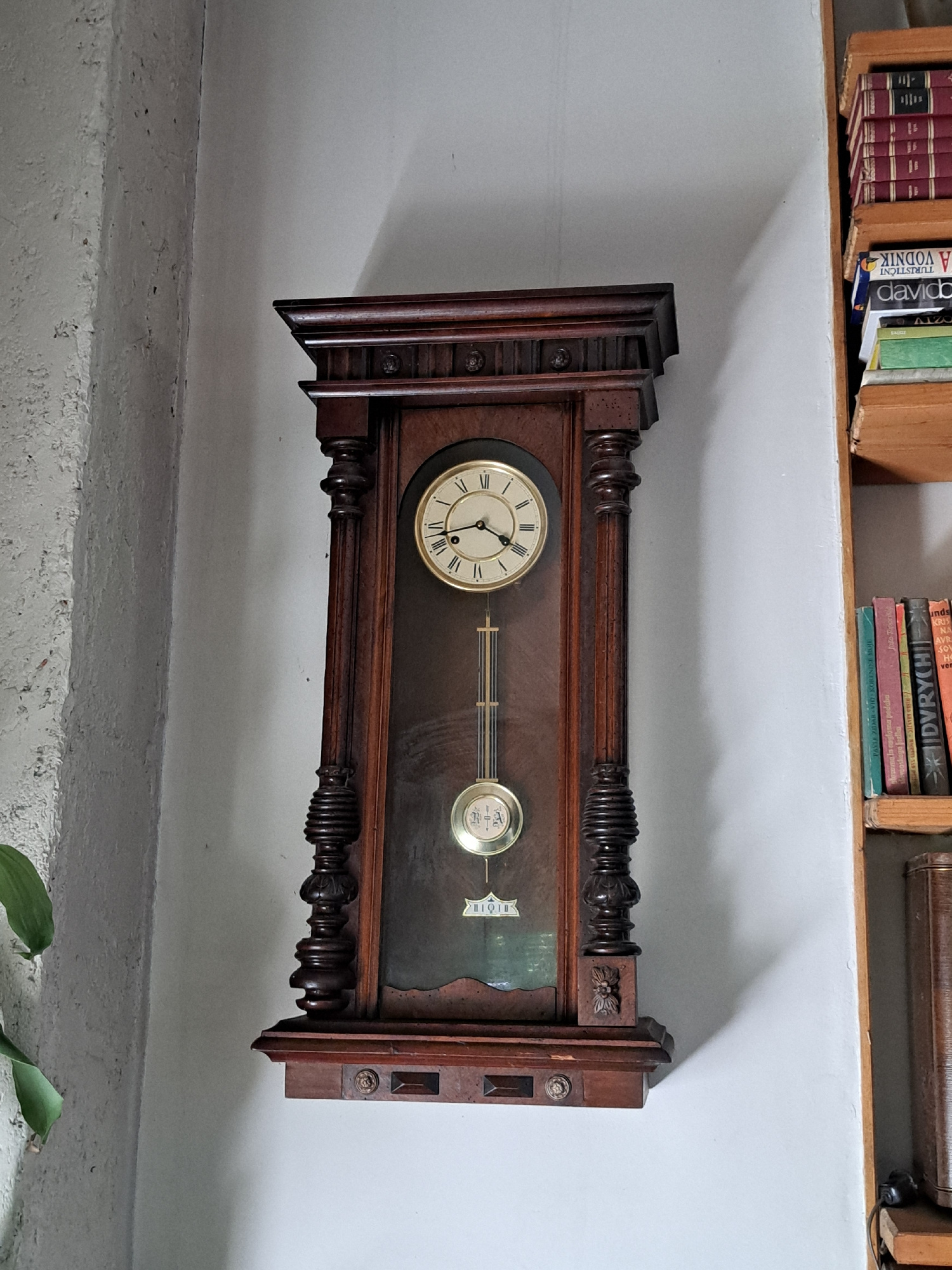 The image shows a pendulum clock mounted on a light-colored wall, with a shelf of books visible above it. The clock is made of dark wood and features a circular face with Roman numerals and a swinging pendulum. The books on the shelf are of various sizes and colors, with some titles partially visible in the Cyrillic alphabet. The overall scene suggests a domestic setting, possibly a living room or study, with an emphasis on traditional decor. The lighting appears to be natural, casting a soft shadow on the wall behind the clock. No people are visible in the image.
