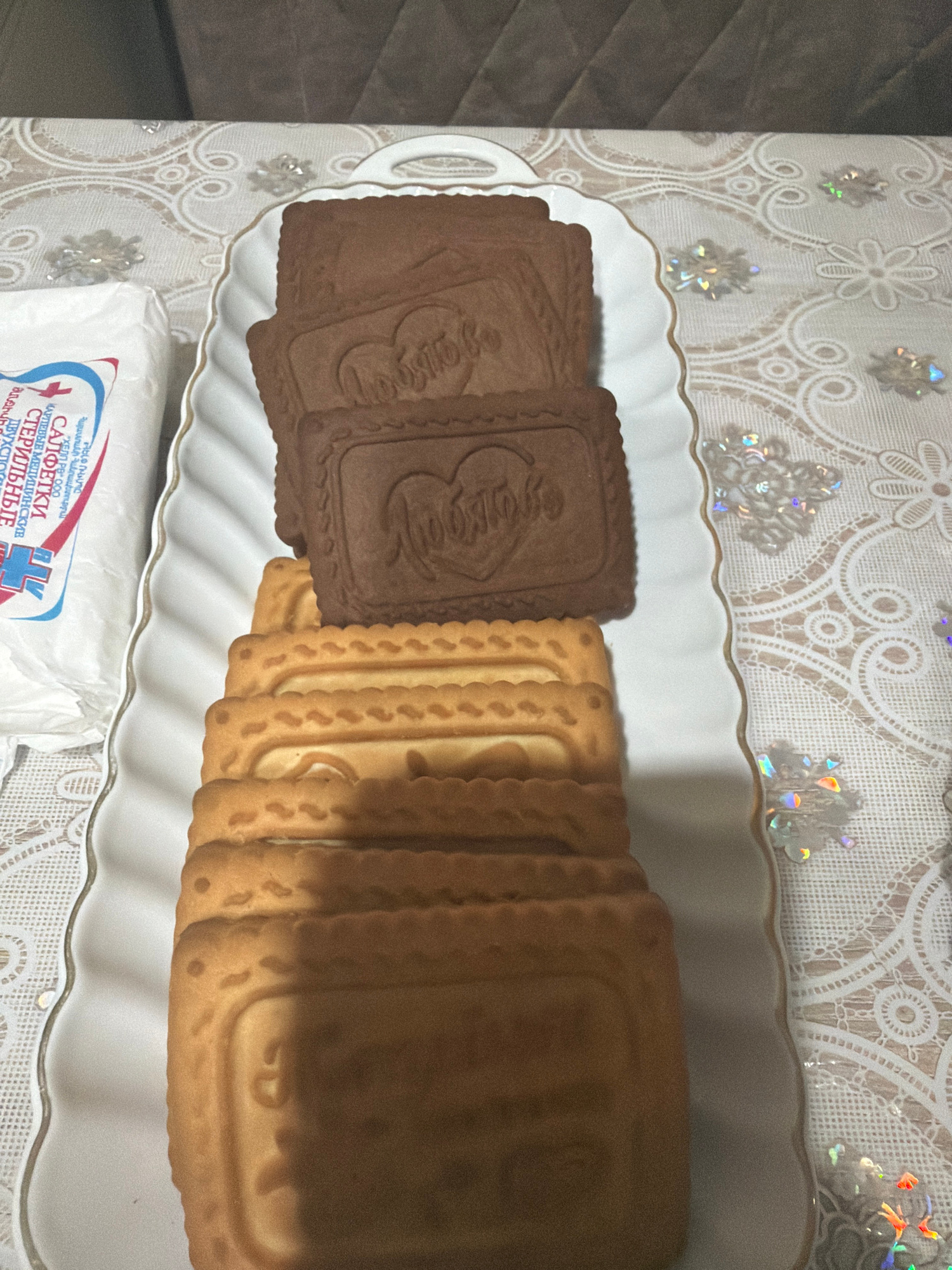 A white rectangular serving platter filled with cookies sits on a patterned tablecloth. The platter contains several layers of rectangular cookies, some light brown and some dark brown. The dark brown cookies have a heart-shaped design with Cyrillic lettering on them. To the left of the platter is a white package with red and blue writing. The tablecloth has a delicate white lace-like pattern with some iridescent snowflake-shaped embellishments scattered across it. The background shows a textured brown surface, possibly a couch. The image appears to be taken indoors, likely in a home setting. The specific location in Yerevan, Armenia cannot be determined from the provided image.