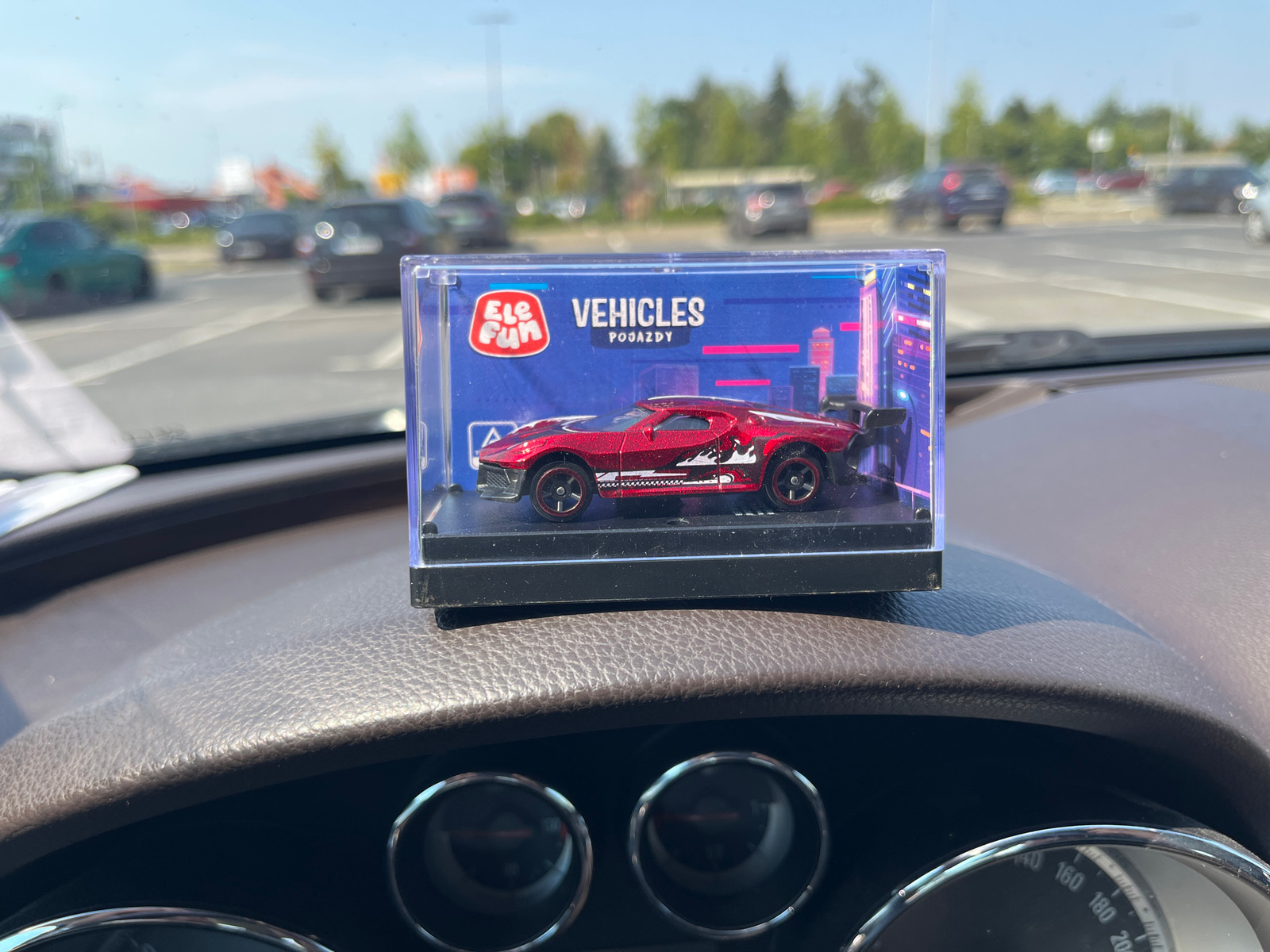The media file is a medium shot of a toy car displayed on a car's dashboard. The toy car is a red, metallic sports car with black and red accents, housed in a clear plastic display case. The case has a blue background with graphics of a cityscape and the text "Ele Fun VEHICLES POJAZDY". The dashboard is dark brown and textured, with visible instrument cluster dials.
In the background, through the car's windshield, a sunny outdoor scene is visible. It appears to be a parking lot or a street with several cars parked and driving. Trees and buildings are discernible in the distance. The sky is clear and blue, suggesting it is daytime with good weather. The location is identified as Piaseczno, Poland. No people are visible in the image. The overall impression is of a personal snapshot, possibly taken by someone who has just purchased or is showcasing the toy car.