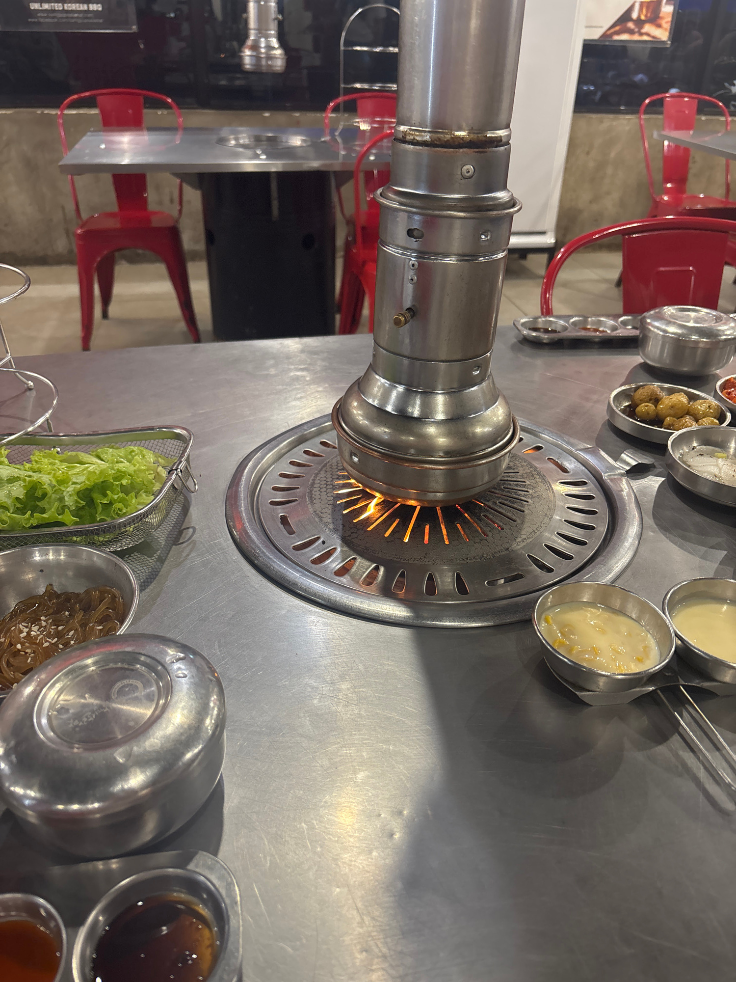 The image shows a close-up view of a Korean BBQ grill setup in a restaurant. The central focus is the circular grill with visible flames beneath a metal grate. Above the grill, a stainless steel ventilation hood is prominently featured. Surrounding the grill are various small metal bowls containing food items. These include what appear to be small potatoes, corn kernels in a creamy sauce, and other unidentifiable food items. To the right of the grill, a metal basket holds fresh green lettuce. Another bowl contains what looks like stir-fried noodles with sesame seeds. In the background, glimpses of red chairs and tables suggest the dining area of a restaurant. The overall impression is a prepared setting for a Korean BBQ meal. The lighting suggests an indoor environment, likely during operating hours. A portion of a sign is visible on the lower left, with text that includes "UNLIMITED KOREAN BBQ." The scene is set in a restaurant, and the activity is the preparation or imminent start of a Korean BBQ meal.