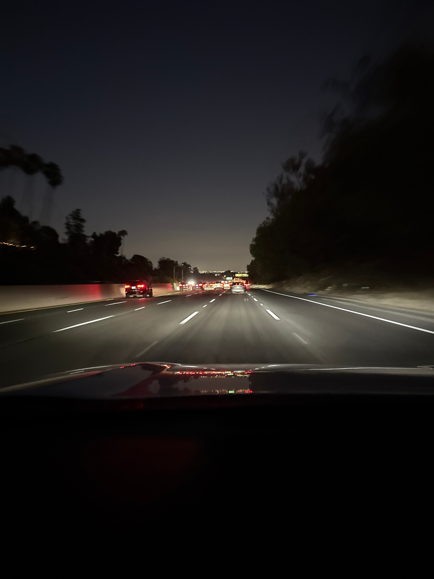 The image depicts a view from inside a vehicle driving on a multi-lane highway at night, likely in Los Angeles, United States of America. The highway has clear lane markings and is illuminated by the headlights of the moving vehicles and streetlights in the distance. Traffic appears to be moderate, with cars visible in multiple lanes ahead. The surrounding environment is dark, with trees and hills visible along the side of the road, suggesting a location with natural elements on the outskirts of the city. The sky is dark, indicating nighttime. The overall scene conveys a sense of travel and urban movement during the evening.