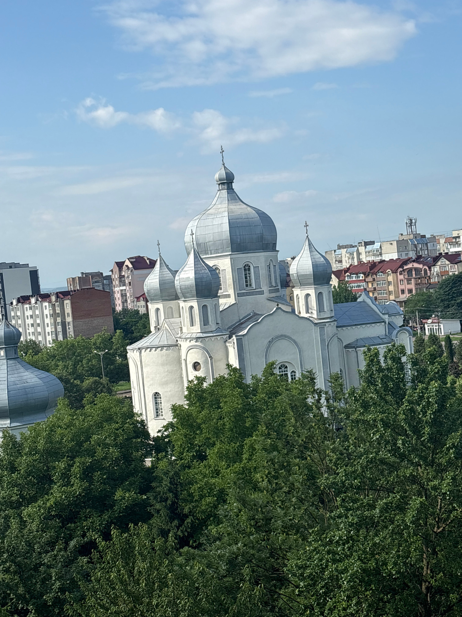 An exterior, high-angle, medium shot captures a large, white, Eastern Orthodox church with multiple silver domes, nestled amongst lush green trees, in the city of Kalush, Ukraine. The church features a prominent central dome topped with a cross, and several smaller domes on its subsidiary structures, also adorned with crosses. The building's architectural style suggests a traditional design with arched windows and ornate detailing. In the background, a cityscape of multi-story buildings, varying in color and style, is visible under a partly cloudy, pale blue sky. The foreground is dominated by the dense foliage of mature trees, partially obscuring the lower sections of the church and the urban landscape. No people are visible in the frame. The lighting suggests it is daytime, with the sun casting a gentle illumination on the scene. The overall impression is one of a historic religious landmark situated within a contemporary urban environment.
