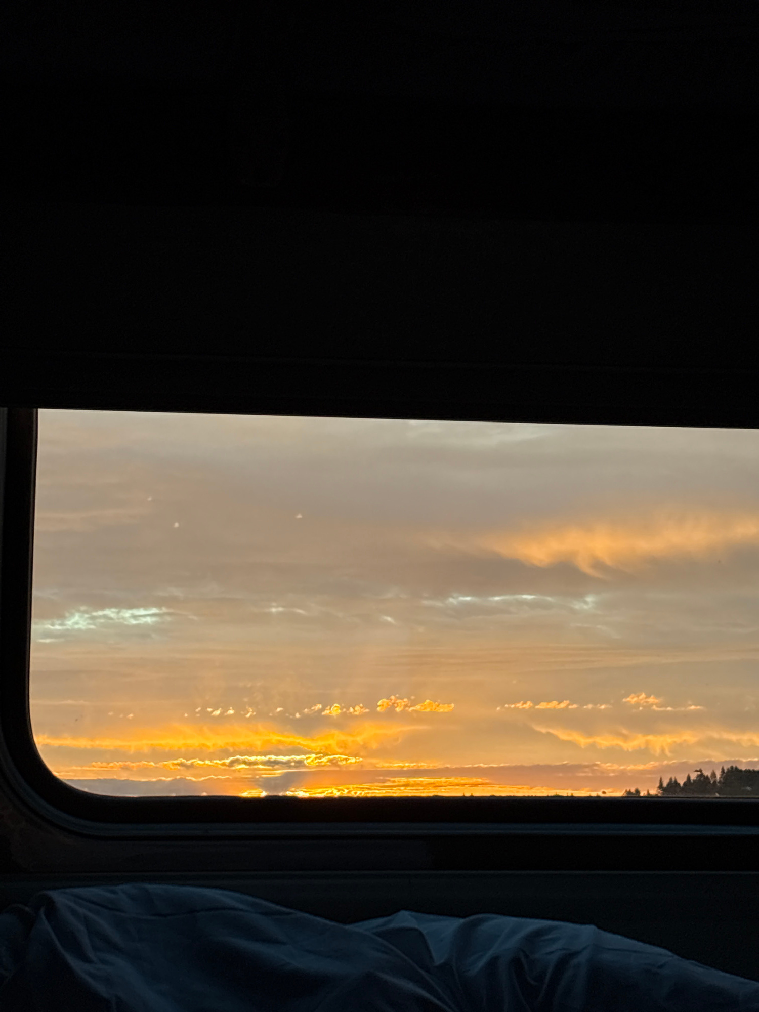 The image captures a sunset viewed through a window. The sky is filled with wispy clouds illuminated by the warm, golden hues of the setting sun, creating a dramatic and serene atmosphere. The window frame, a dark, rounded structure, encloses a portion of the sky, with a dark, solid object to the left and a crumpled blue fabric on the right, possibly part of a curtain or bedding. The scene suggests a moment of quiet observation, perhaps from a vehicle or a room. The time of day is clearly evening, indicated by the sunset. The weather appears clear aside from the clouds, and the lighting is soft and warm. There are no visible people or text in the image. The location context is Торбино, Russia.