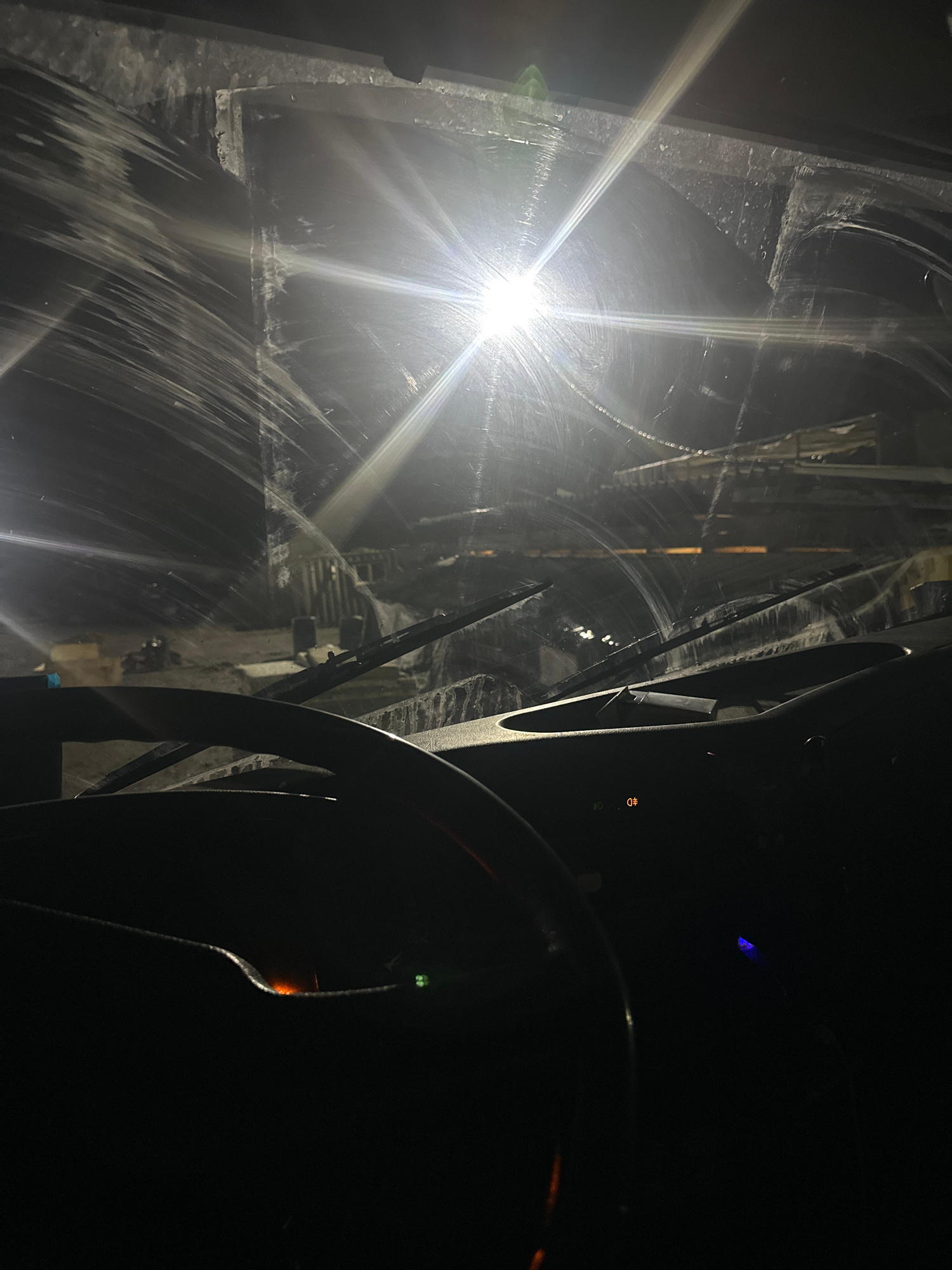 The image provides a view from inside a vehicle, looking through the windshield at night. The windshield is heavily streaked with what appears to be water or dirt, distorting the view of the exterior. A bright light source, possibly headlights or a streetlight, creates a strong glare and lens flare effect, obscuring many details of the external environment. The interior of the vehicle is dark, with the steering wheel visible in the foreground on the left. The dashboard has some illuminated indicators, including what looks like a digital display showing "09", and other unidentifiable lights. The exterior scene is largely indistinct due to the glare and streaks, but suggests an urban or industrial setting with structures and some lighting in the distance. The overall impression is that of driving in challenging conditions at night.