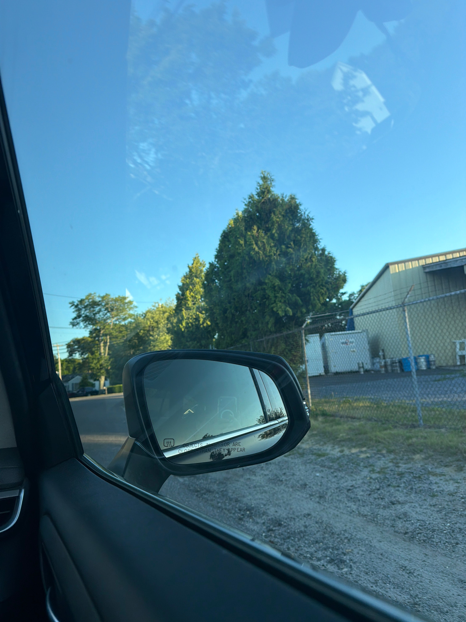 The image captures a view from inside a vehicle, looking out through the driver's side window and reflecting in the side mirror. The sky is a clear, bright blue with some wispy clouds, suggesting a sunny day. In the foreground, the interior of the car is visible, including the dark-colored door panel and a portion of the window frame. The side mirror, a prominent element, reflects the sky, trees, and what appears to be a building or structure.
The scene outside the car reveals a suburban or semi-industrial environment. To the left, a residential street is visible with houses and trees lining it. Further to the right, there is a chain-link fence bordering an area with a light-colored industrial building, several white storage containers, and some blue barrels. A few trees, including what appear to be tall, conical evergreens, are interspersed in the landscape. The ground in the foreground is a gravelly surface. The overall impression is that of a typical day in a mixed-use area, potentially on the outskirts of a town or city. The text "OBJECTS IN MIRROR ARE CLOSER THAN THEY APPEAR" is visible on the side mirror. The location is confirmed as Riverhead, United States of America.