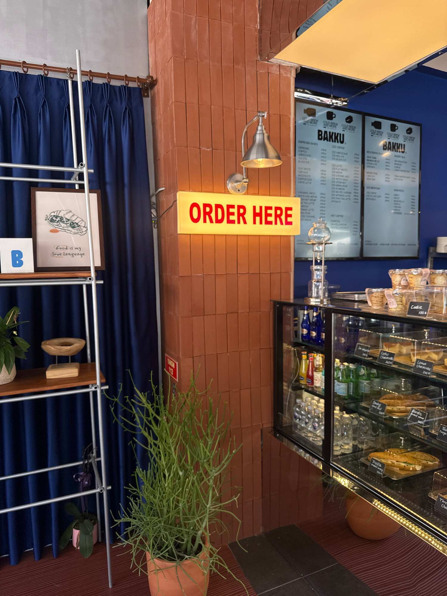 This is a vertically oriented, close-up, eye-level photograph taken inside a cafe. The scene is dominated by a glass display case filled with pastries and drinks. On the left, a menu board is visible, listing various coffee and espresso-based drinks under the "BAKKU" brand. The menu board is illuminated, and a yellow "ORDER HERE" sign is prominently displayed. To the right of the menu, a display case shows items like cookies and cheesecakes. Bottles of beverages and other condiments are visible in the lower section of the display. A green, spiky plant in a terracotta pot sits to the right of the display case. In the foreground, a section of blue velvet curtain is visible, behind which a metal shelving unit holds framed artwork and decorative items. The overall ambiance suggests a vibrant and modern cafe setting, likely during operating hours given the illuminated signage and display items. The decor, including the exposed brick wall and blue curtains, hints at a stylish and contemporary interior. The text visible on the menu boards and display case provides product information and pricing.